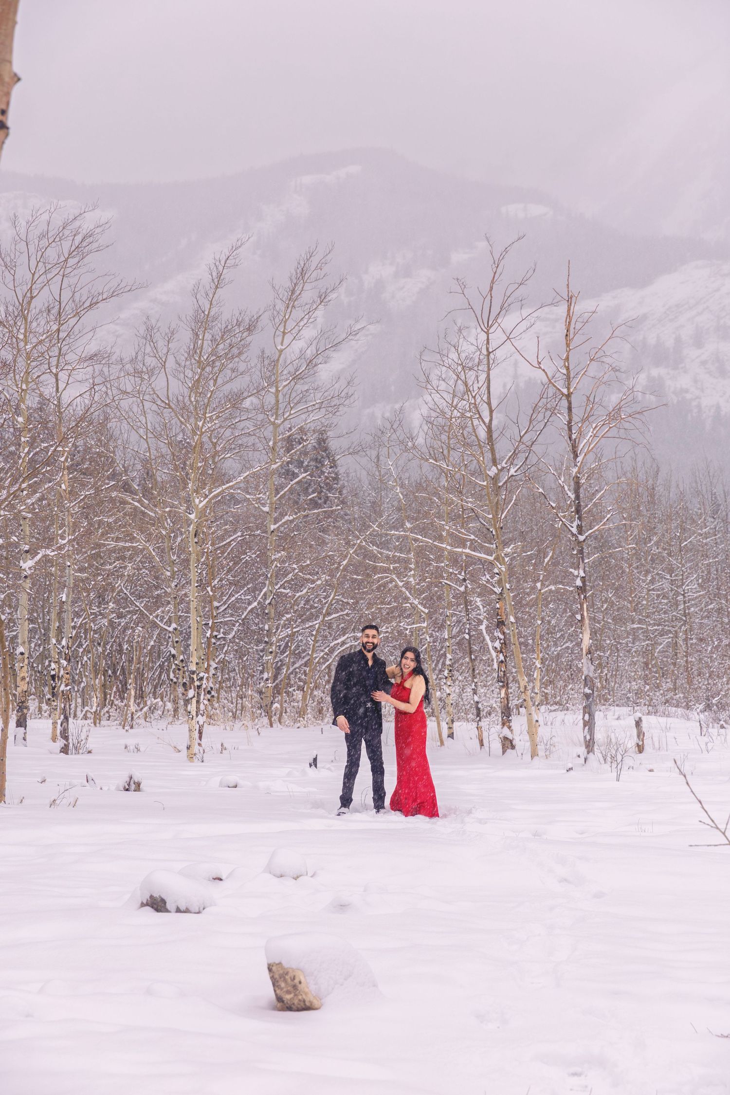 Couple in formal wear embracing in snowy winter landscape with bare aspen trees and mountain backdrop.