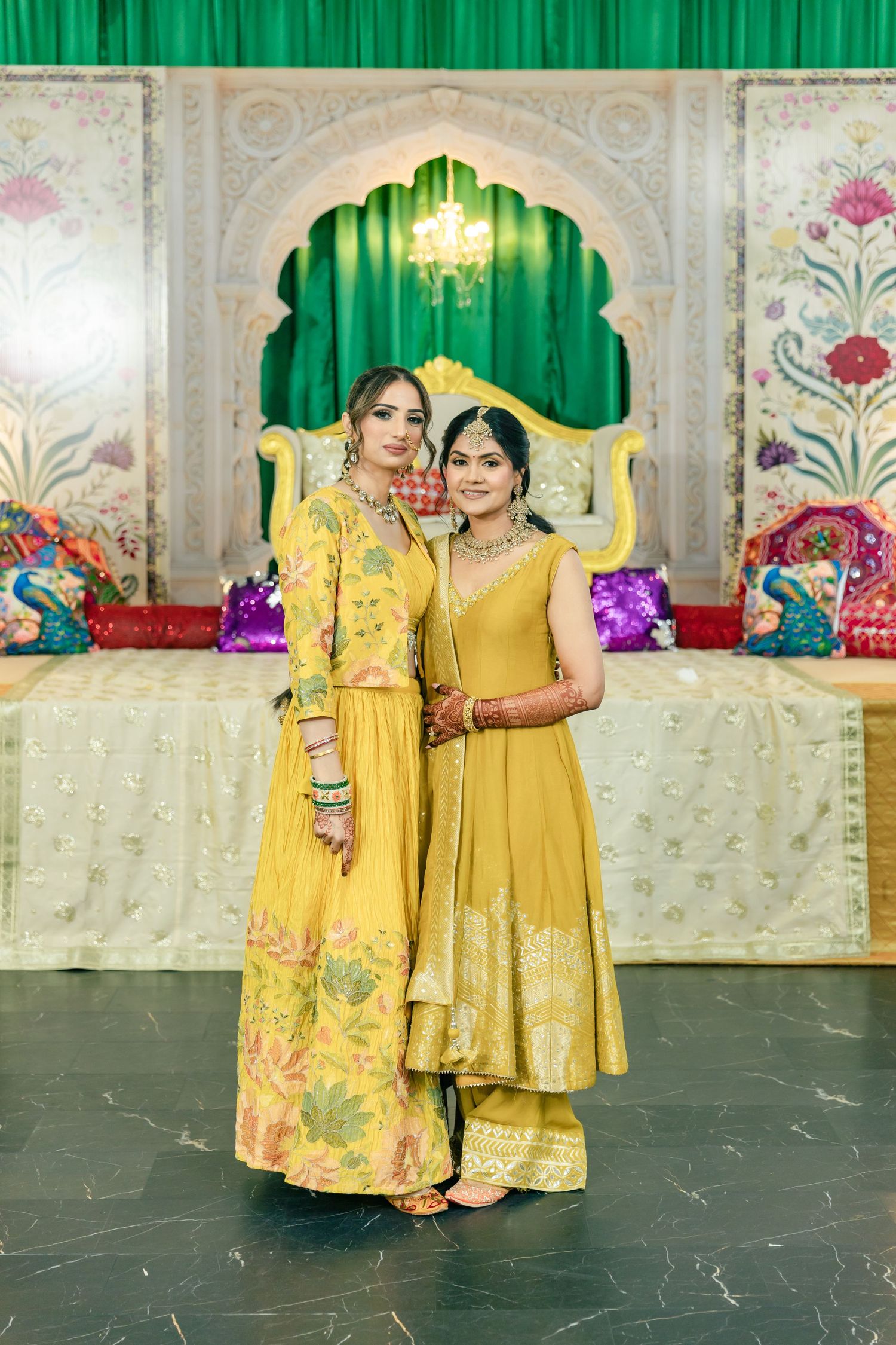 Two people wearing matching yellow traditional Indian outfits pose together against an ornate backdrop with green curtains.