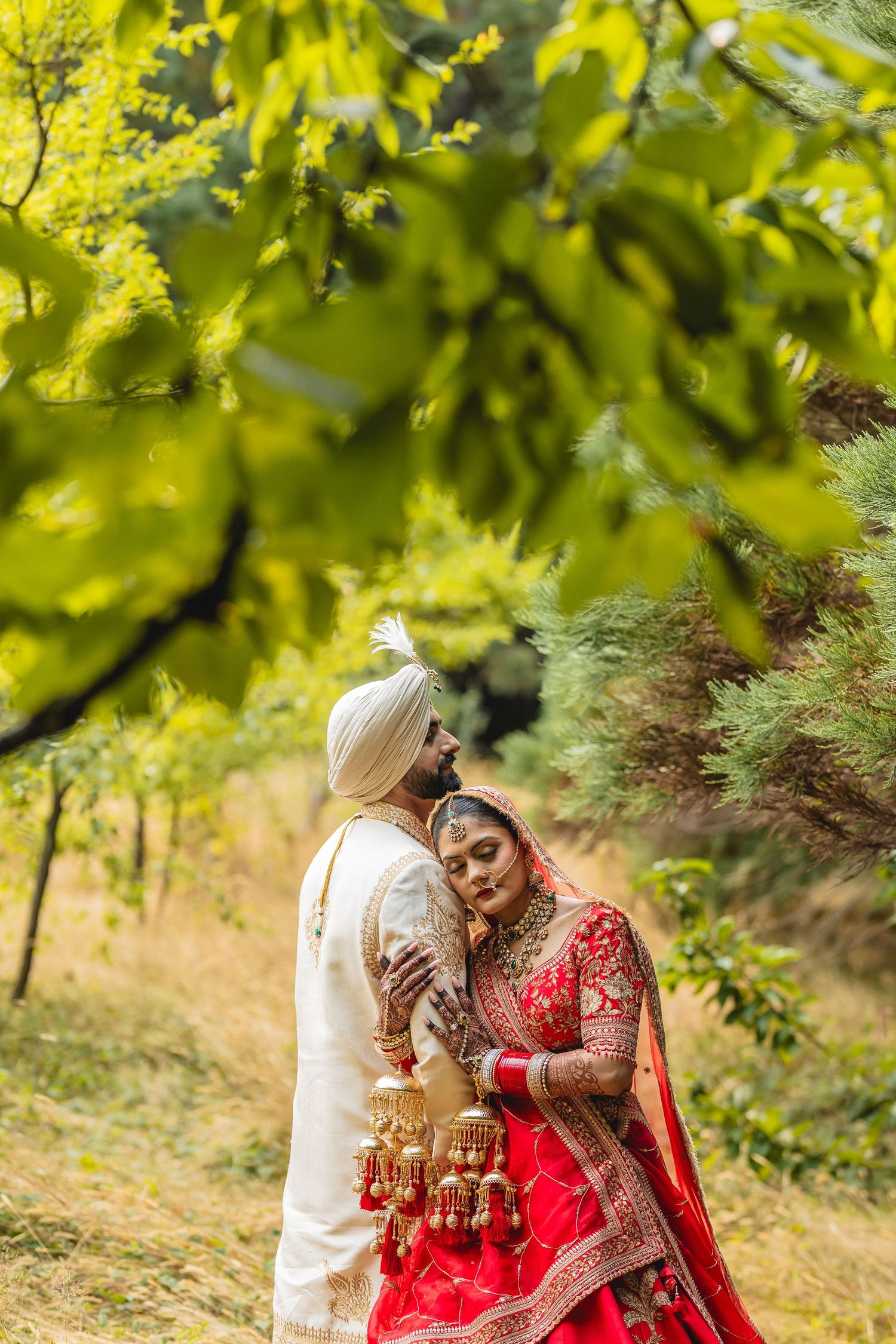 A couple in traditional wedding attire share an intimate moment under sunlit tree branches.