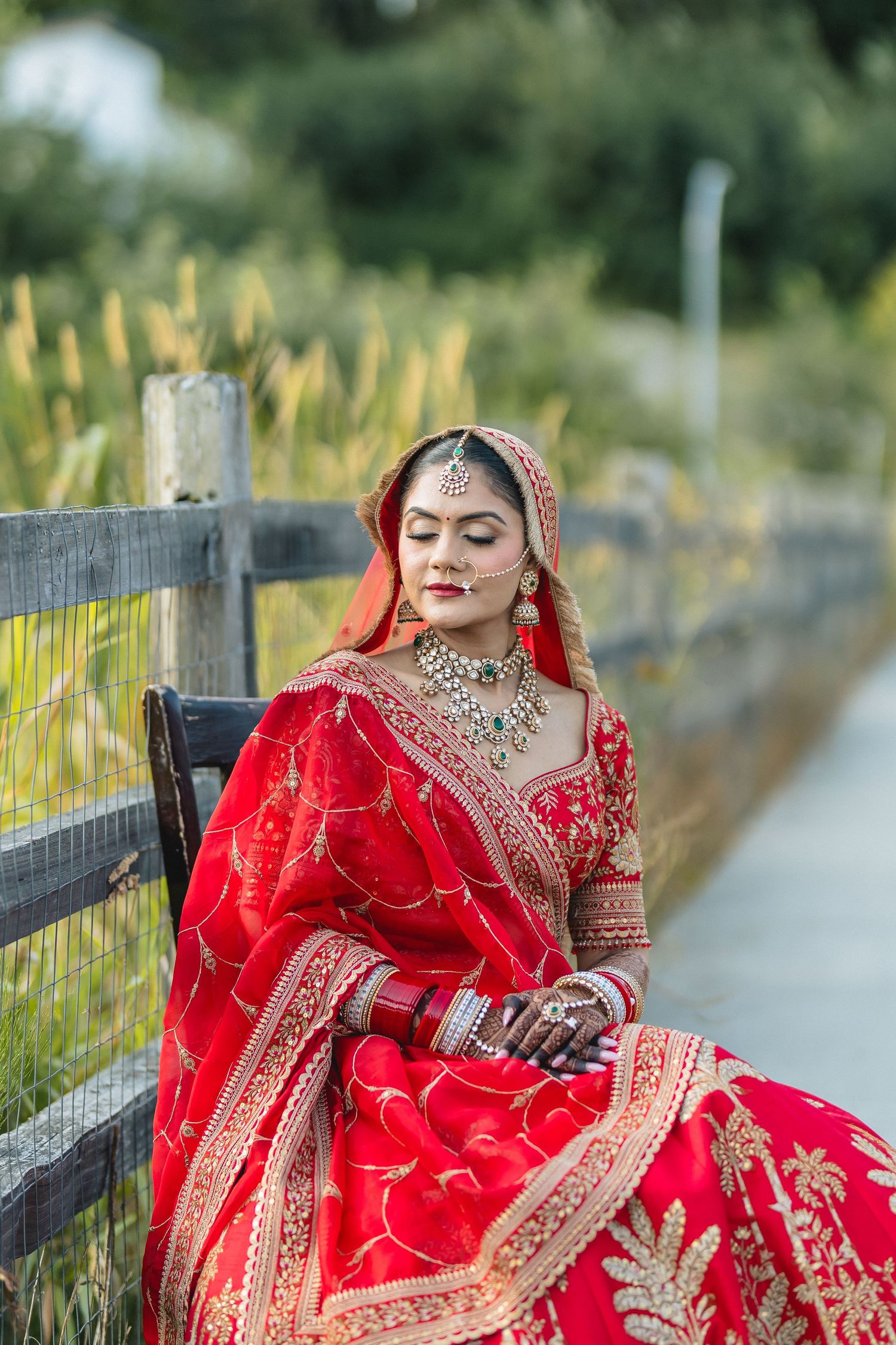Bride in red lehenga poses outdoors against rustic fence and natural backdrop.