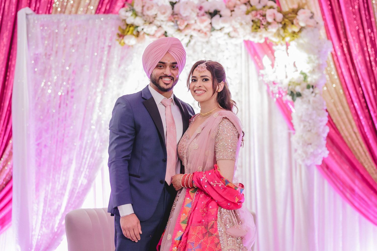 Couple in traditional Punjabi wedding attire posing under pink floral decorations.