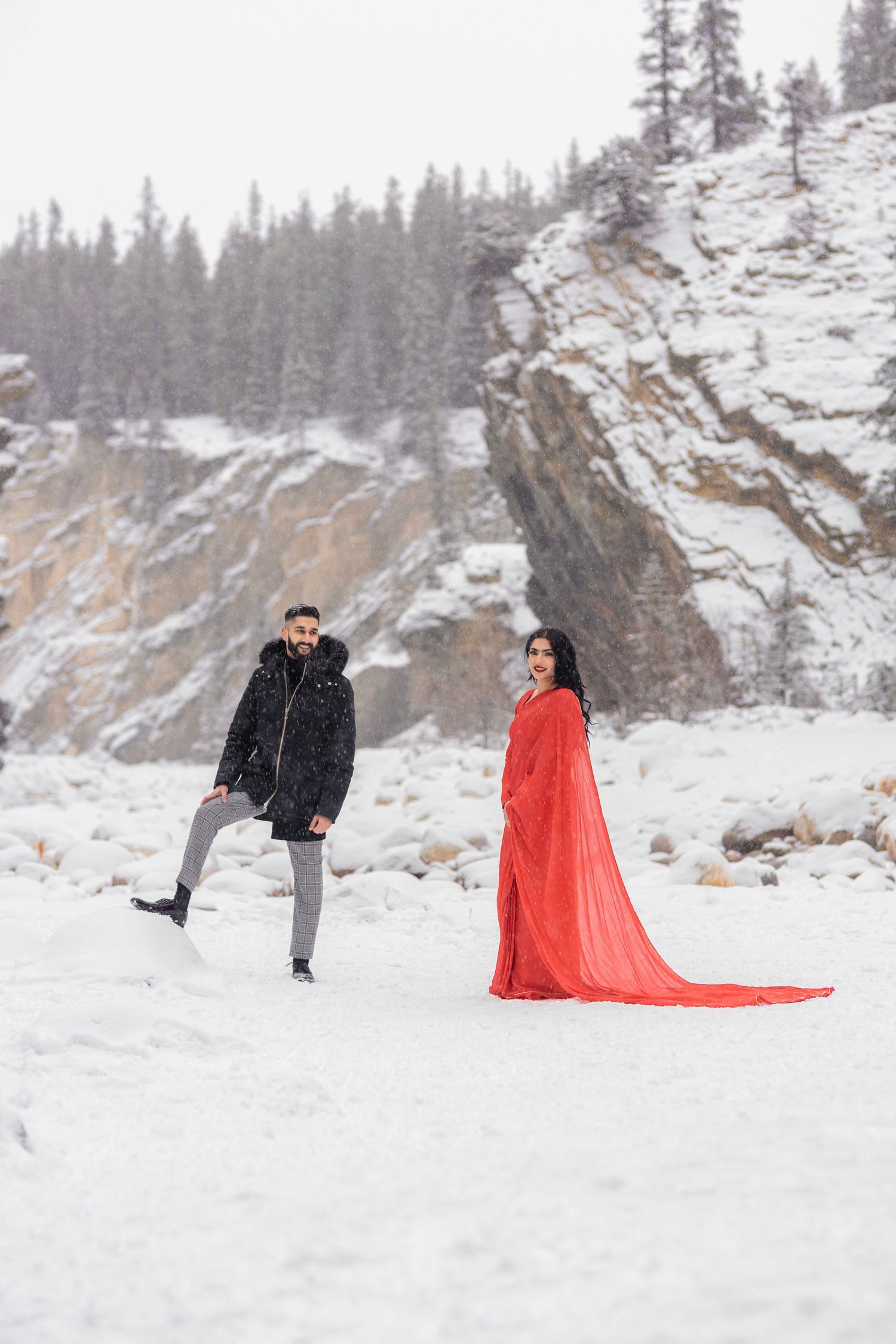 Couple in snowy winter landscape, man in black jacket and woman in flowing red dress with train, rocky cliffs and pine trees behind.
