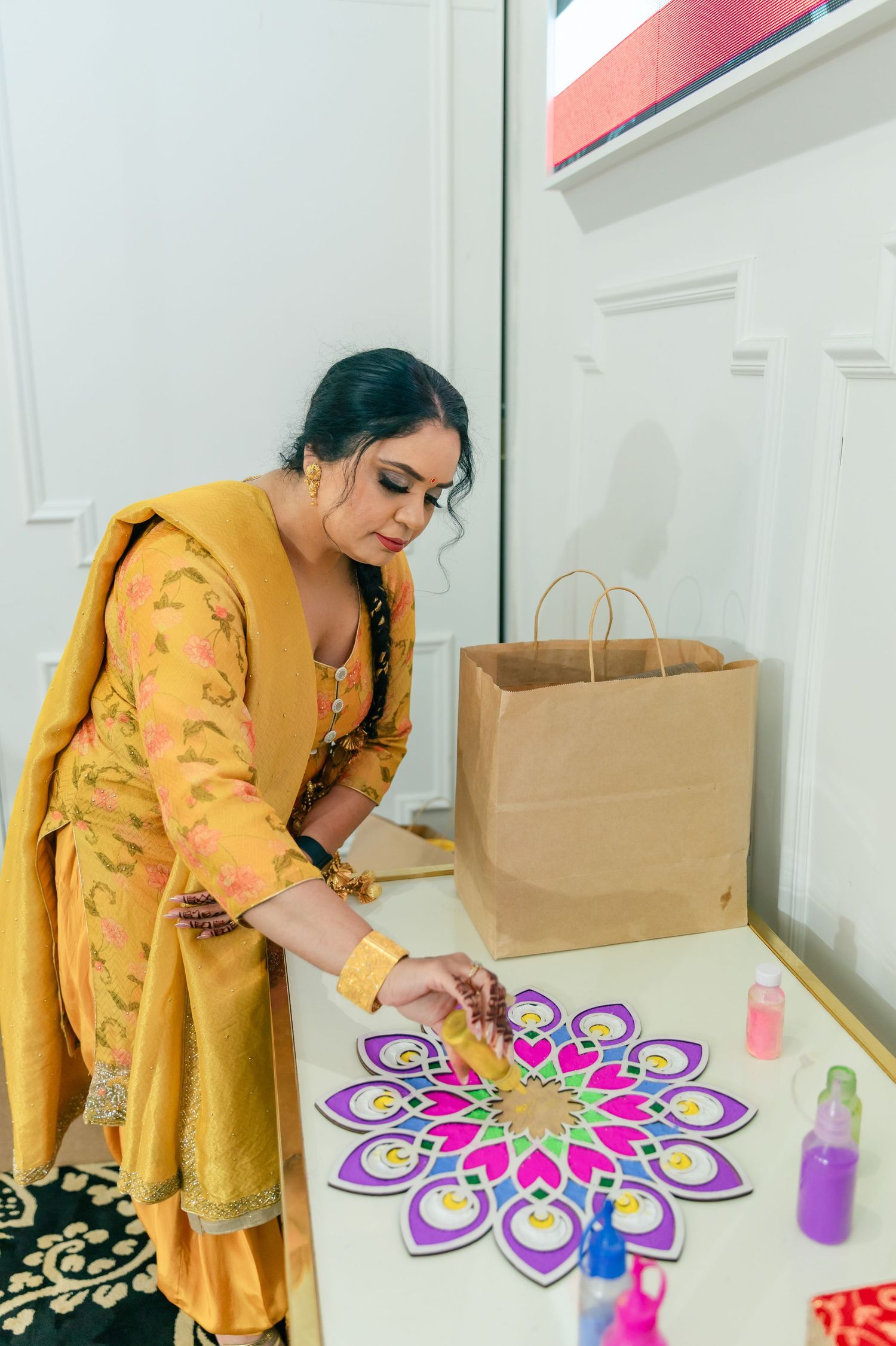 A person in yellow traditional dress creates colorful rangoli art patterns on a white surface.