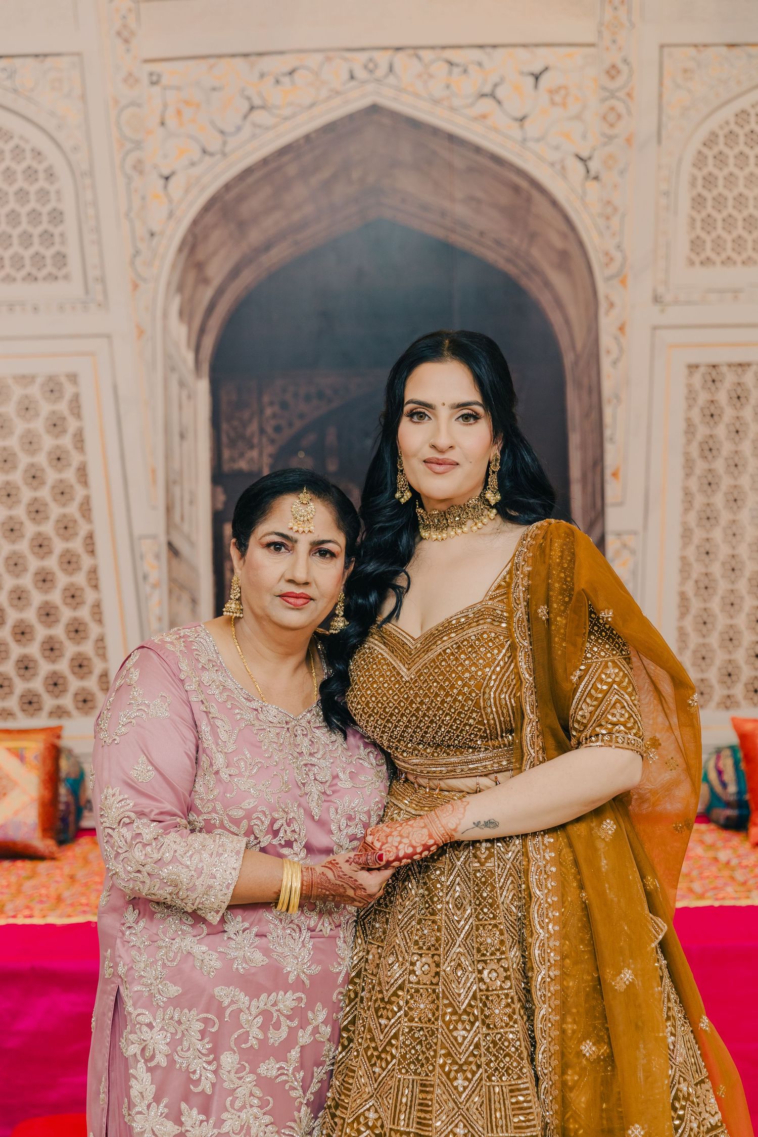 Two women in ornate Indian outfits pose together in front of intricate archway architecture.