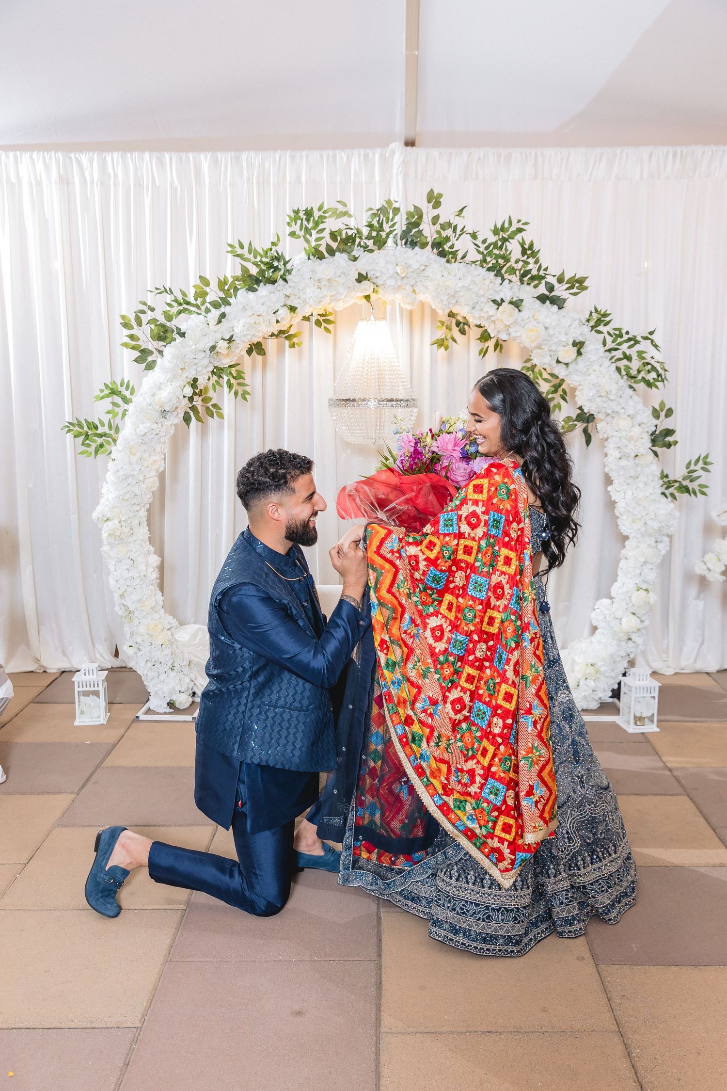 A couple in traditional Indian attire poses together in front of a floral wedding arch with white draping. Captured by Abby Image Studio