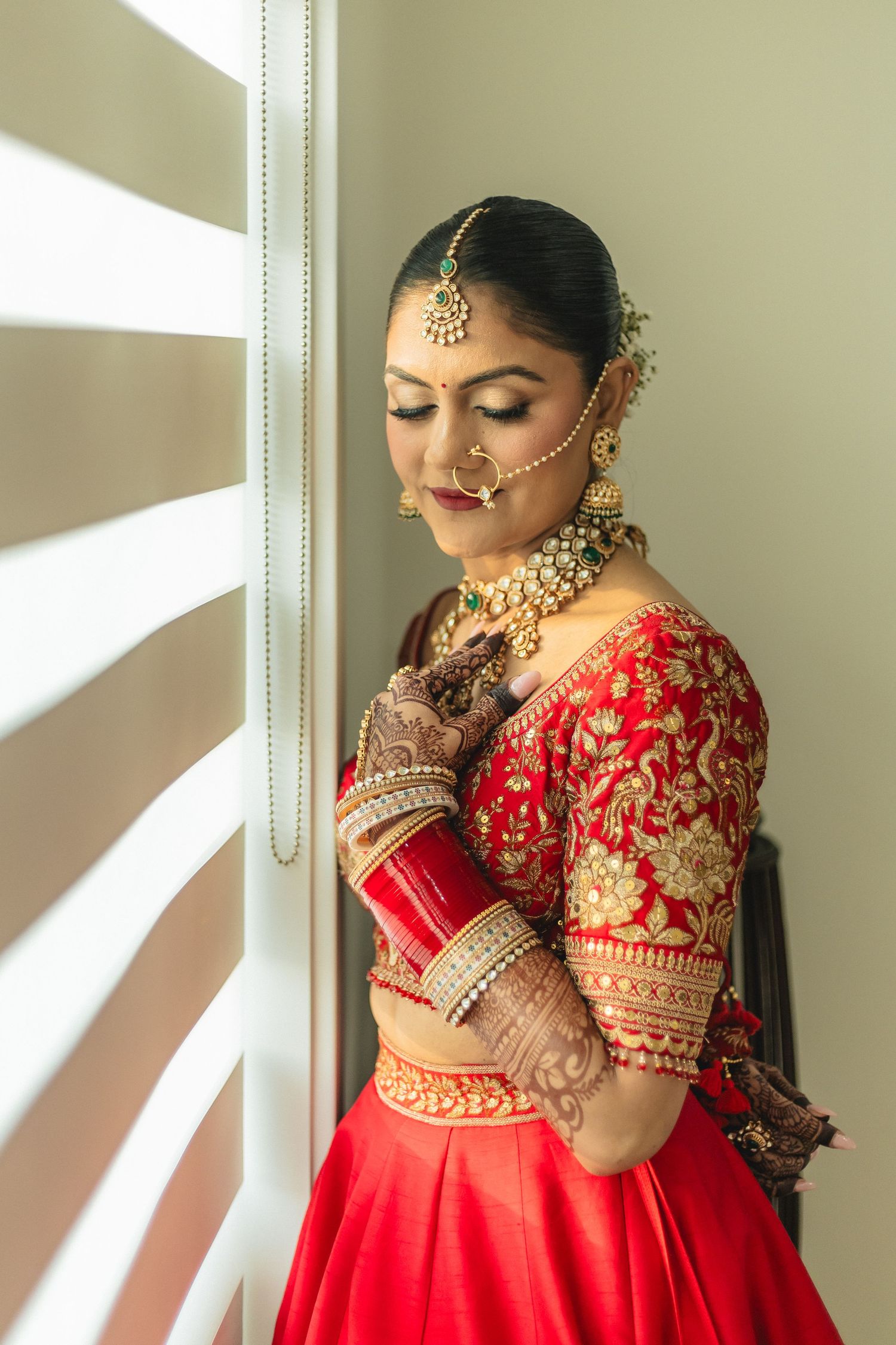 A bride in a traditional red and gold embroidered lehenga poses near a window with natural light.