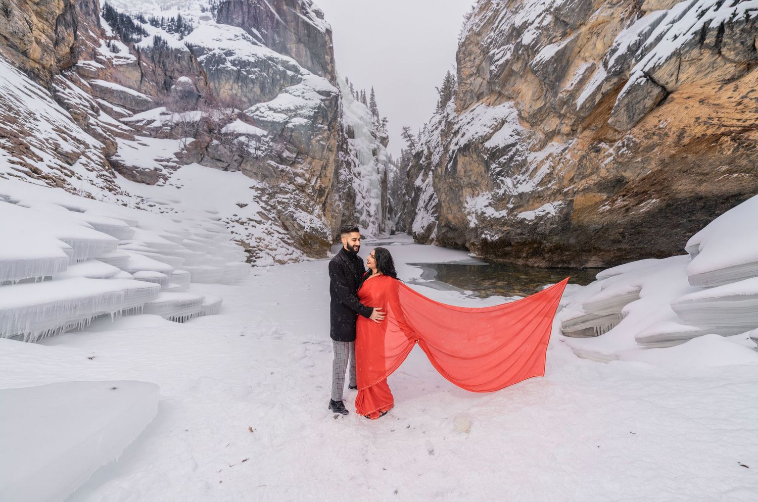 Couple in red dress posing in snowy canyon with frozen waterfall and dramatic rocky cliffs in winter.