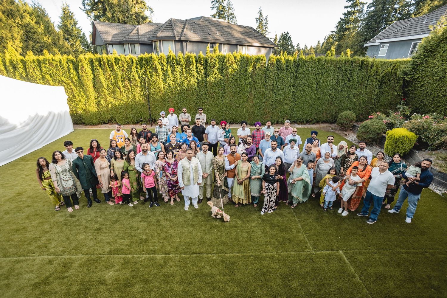 Large outdoor group photo of wedding guests arranged in rows on a sunny lawn with hedges.