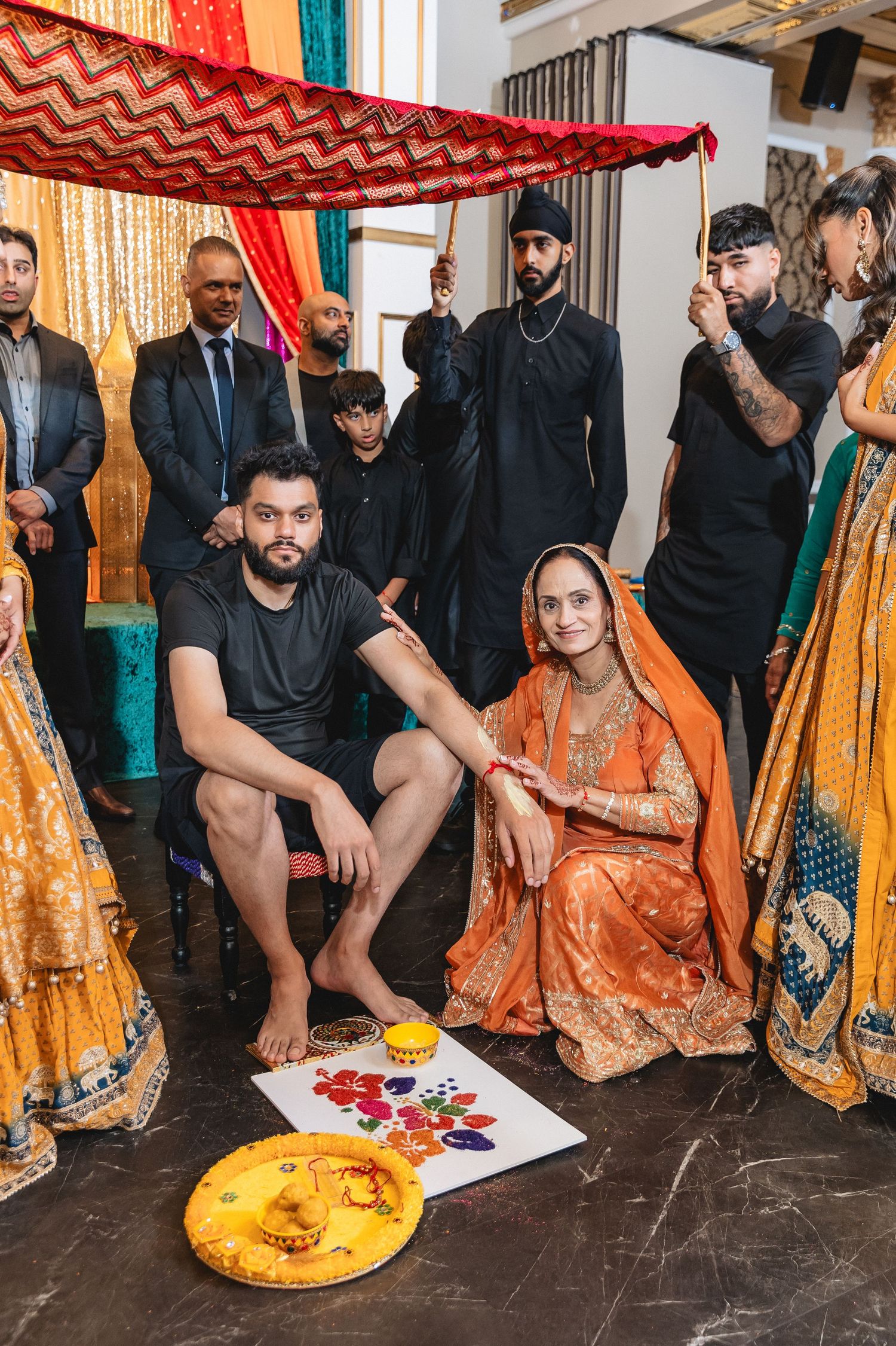 Traditional Indian wedding ceremony guests sit on floor wearing colorful orange and black attire during cultural celebration.