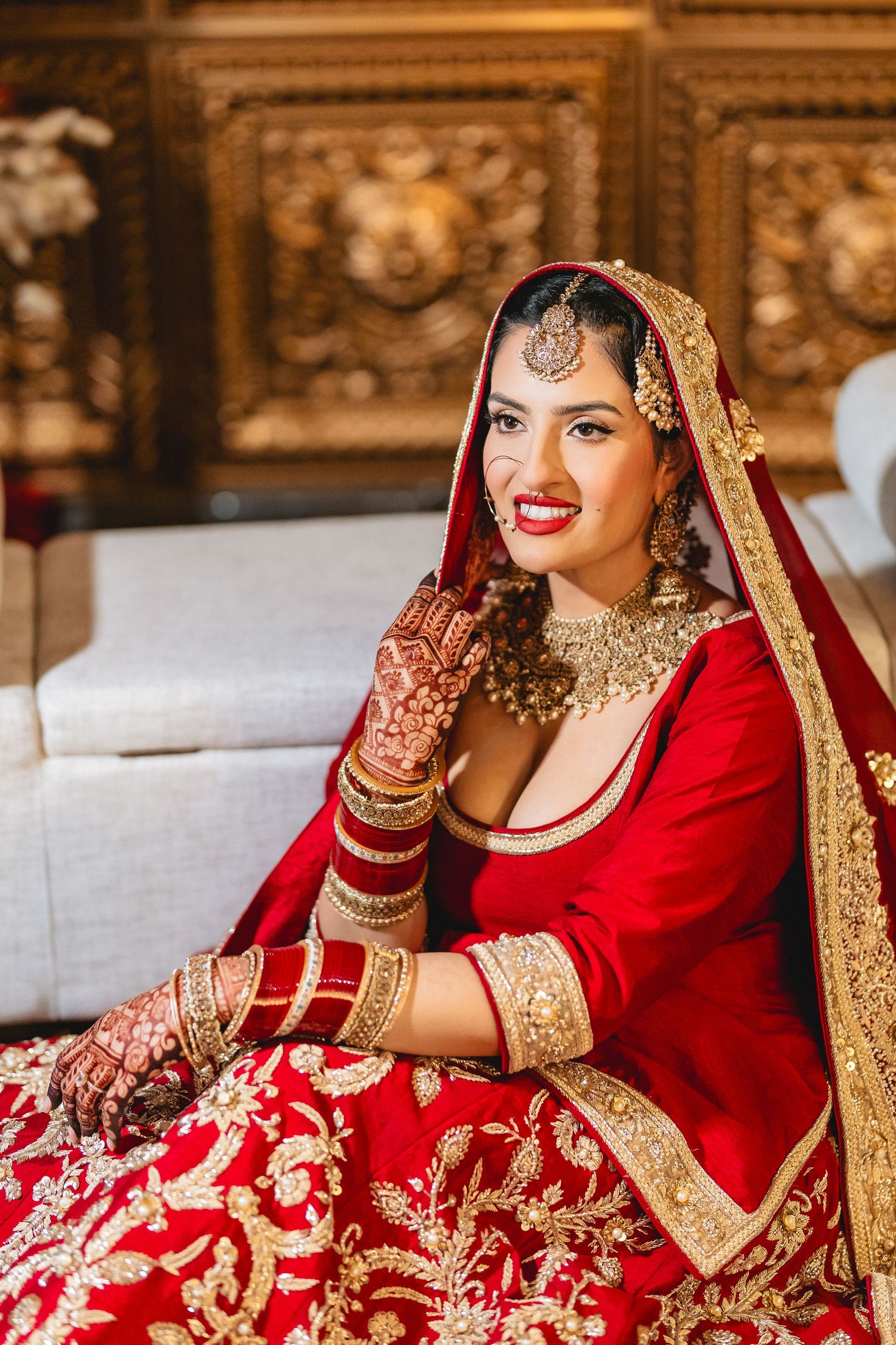 Bride wearing ornate red and gold traditional Indian wedding lehenga poses elegantly on couch.