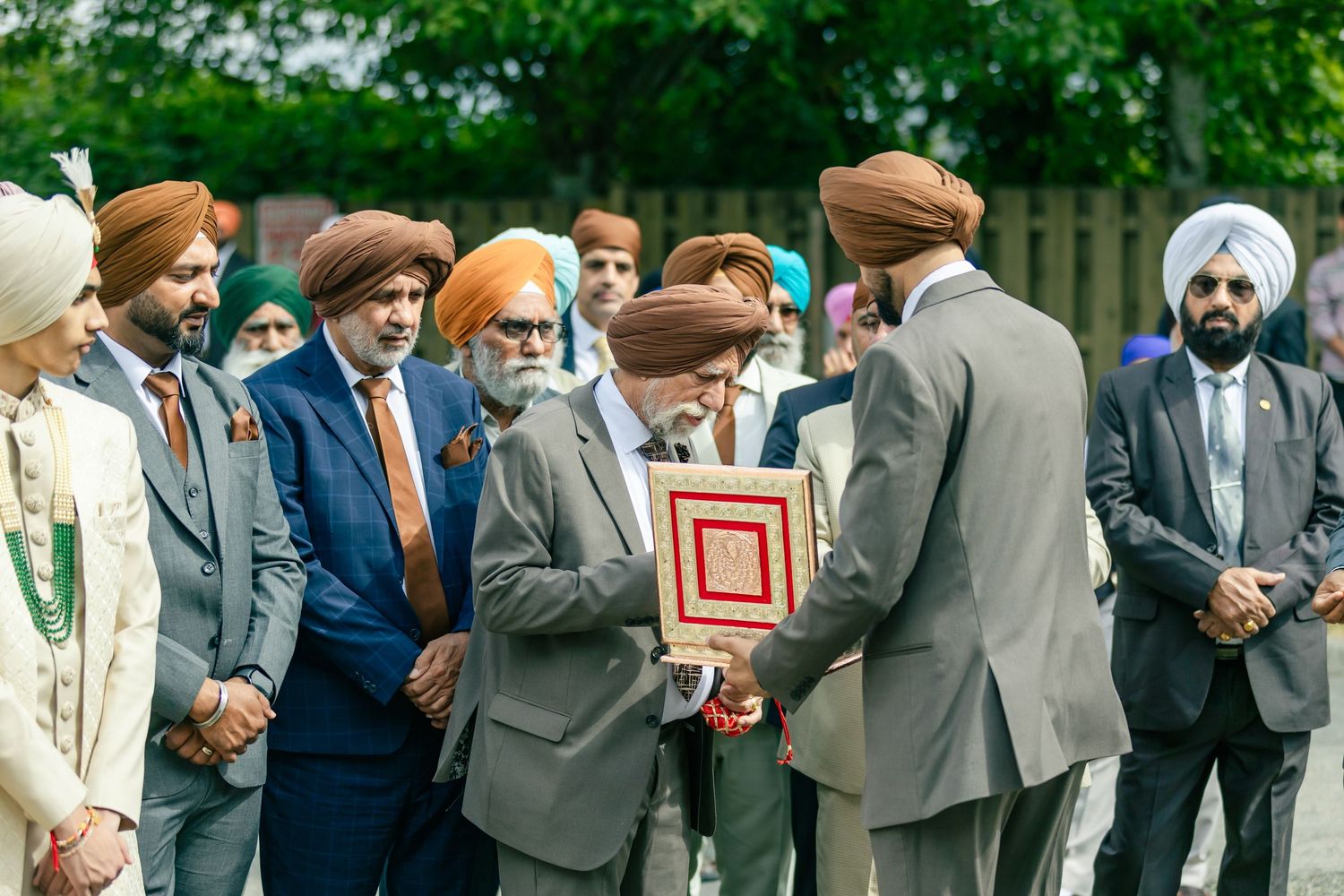 A large group of wedding guests in suits and formal wear stand together outdoors.