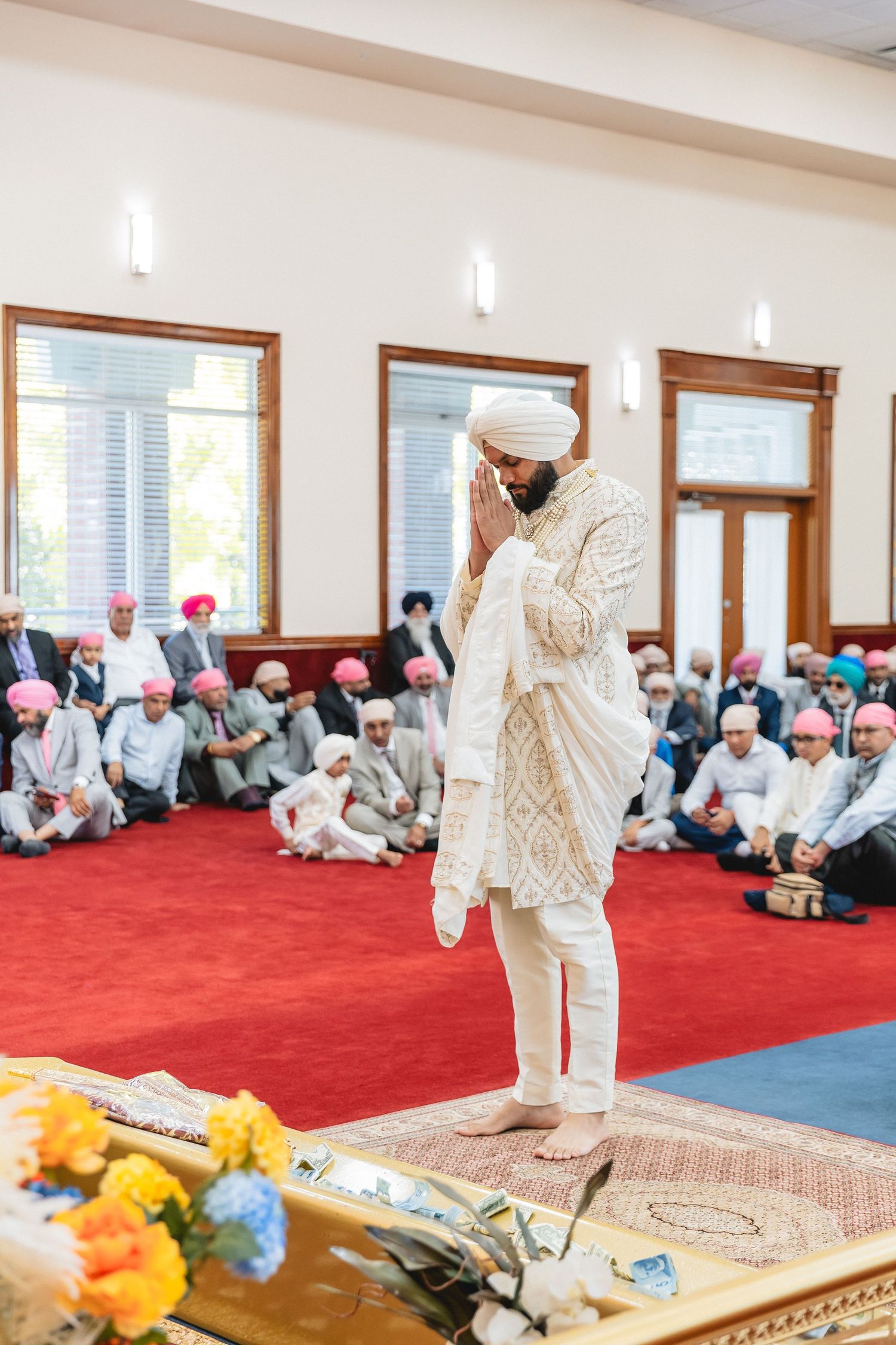 Traditional Sikh wedding ceremony taking place in a temple with red carpeting and prayer mats.