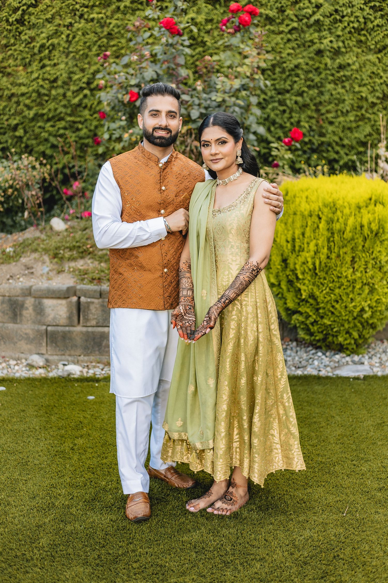 Couple in traditional Indian attire posing in a garden with red flowers and green shrubs.