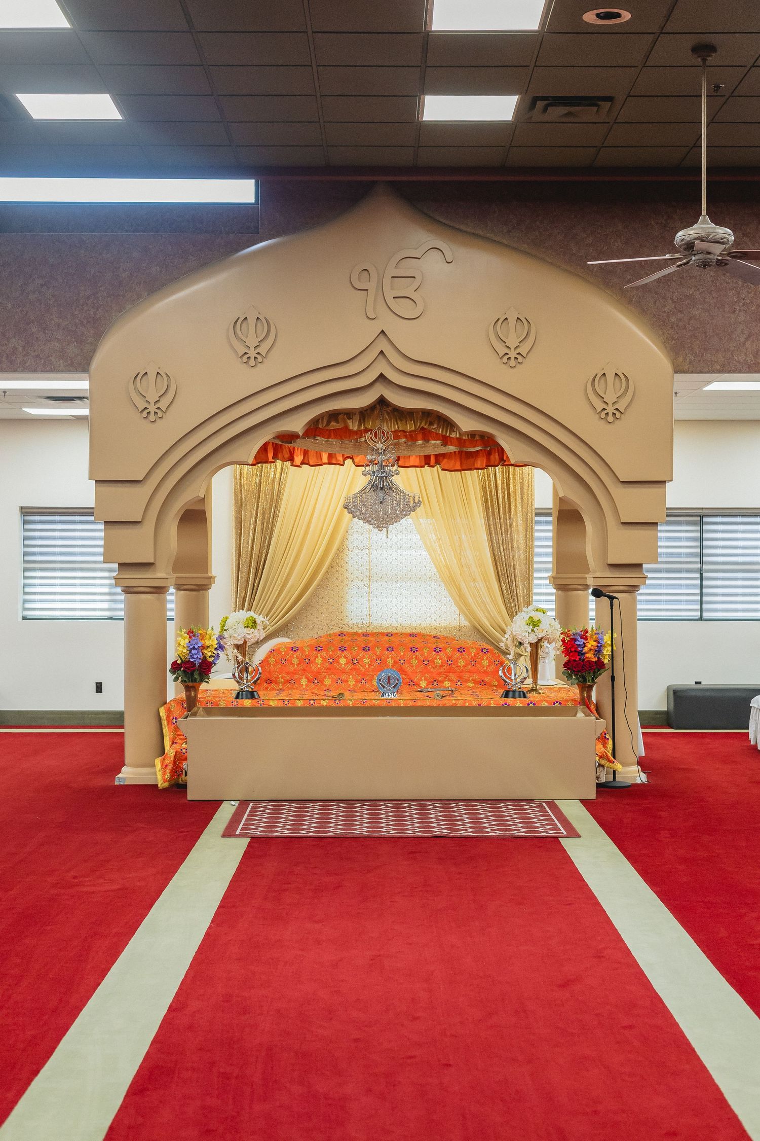 Sikh temple interior with red carpet and ornate stage featuring traditional architectural elements and golden curtains.