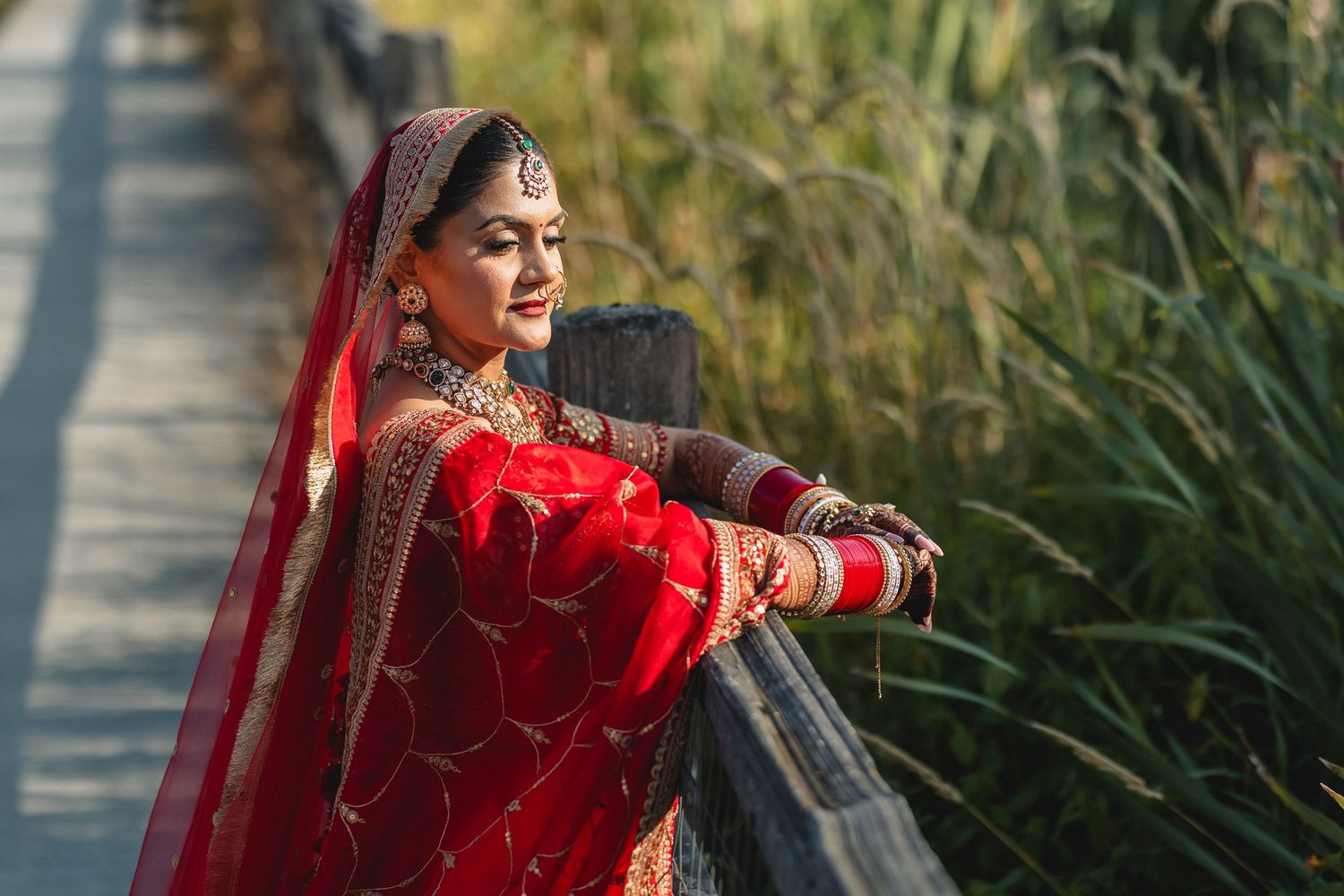 Bride in red traditional attire posing outdoors against tall grass during golden hour.