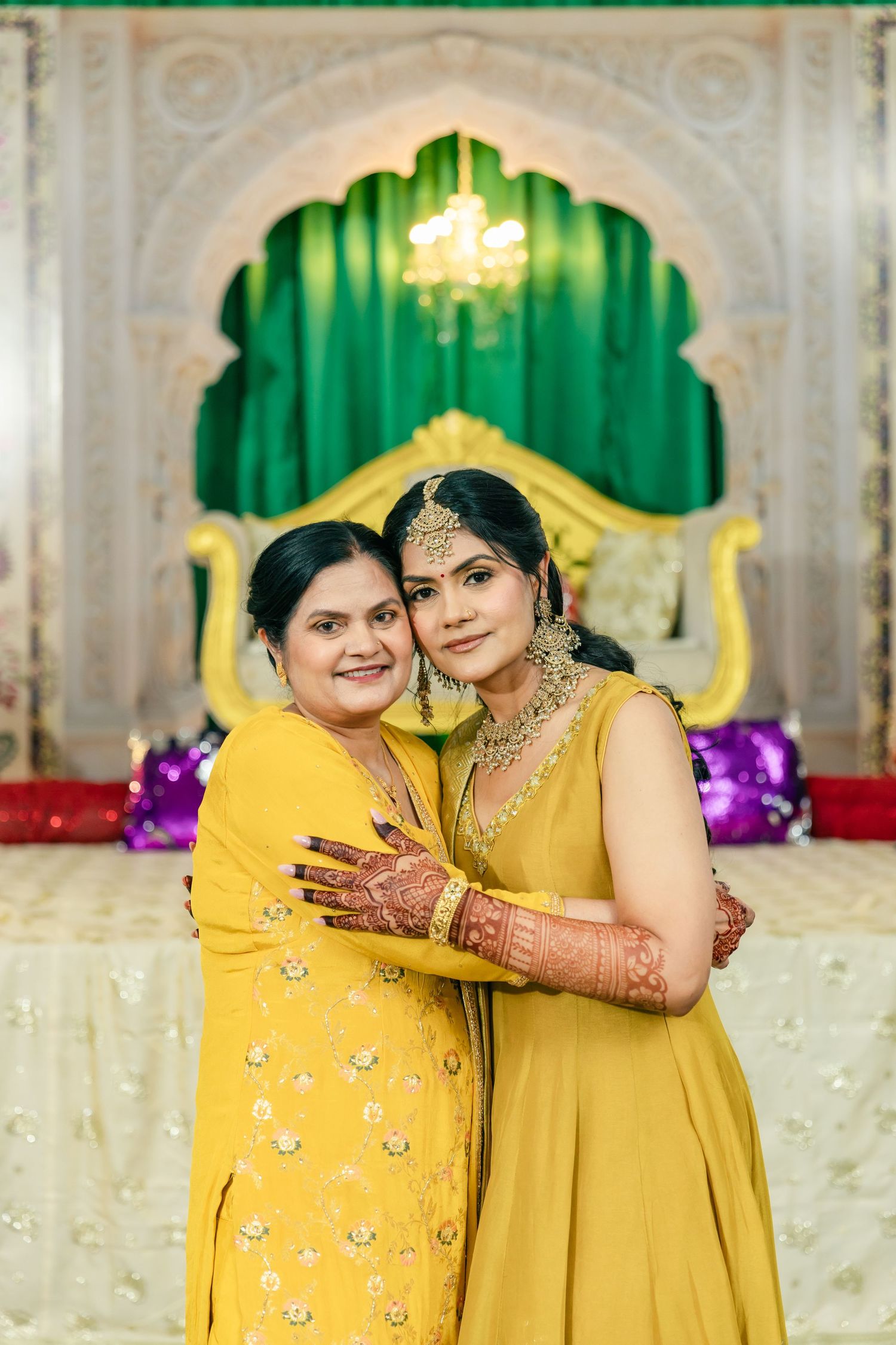 Two guests in yellow traditional Indian outfits pose together at a decorated venue with green backdrop.