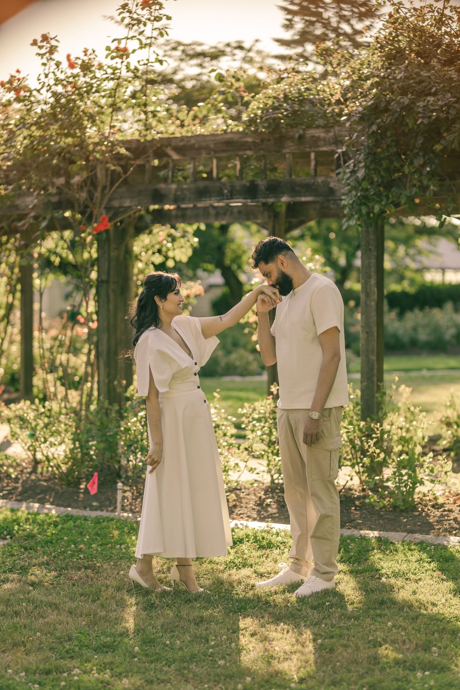 A couple in all-white outfits dances playfully in a sunlit garden setting during golden hour.