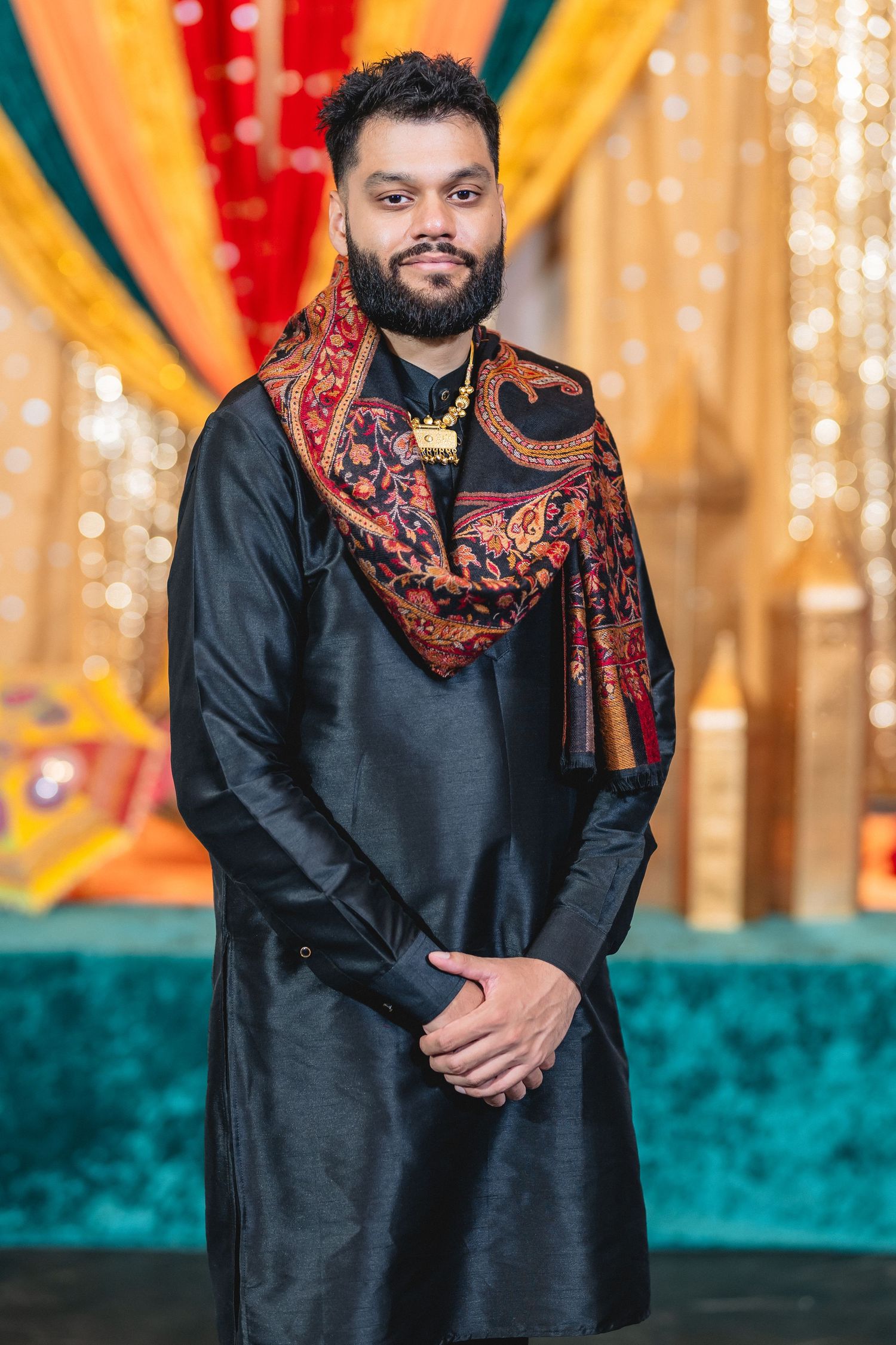 A person in a black traditional outfit with an ornate red and gold scarf poses against a colorful backdrop.
