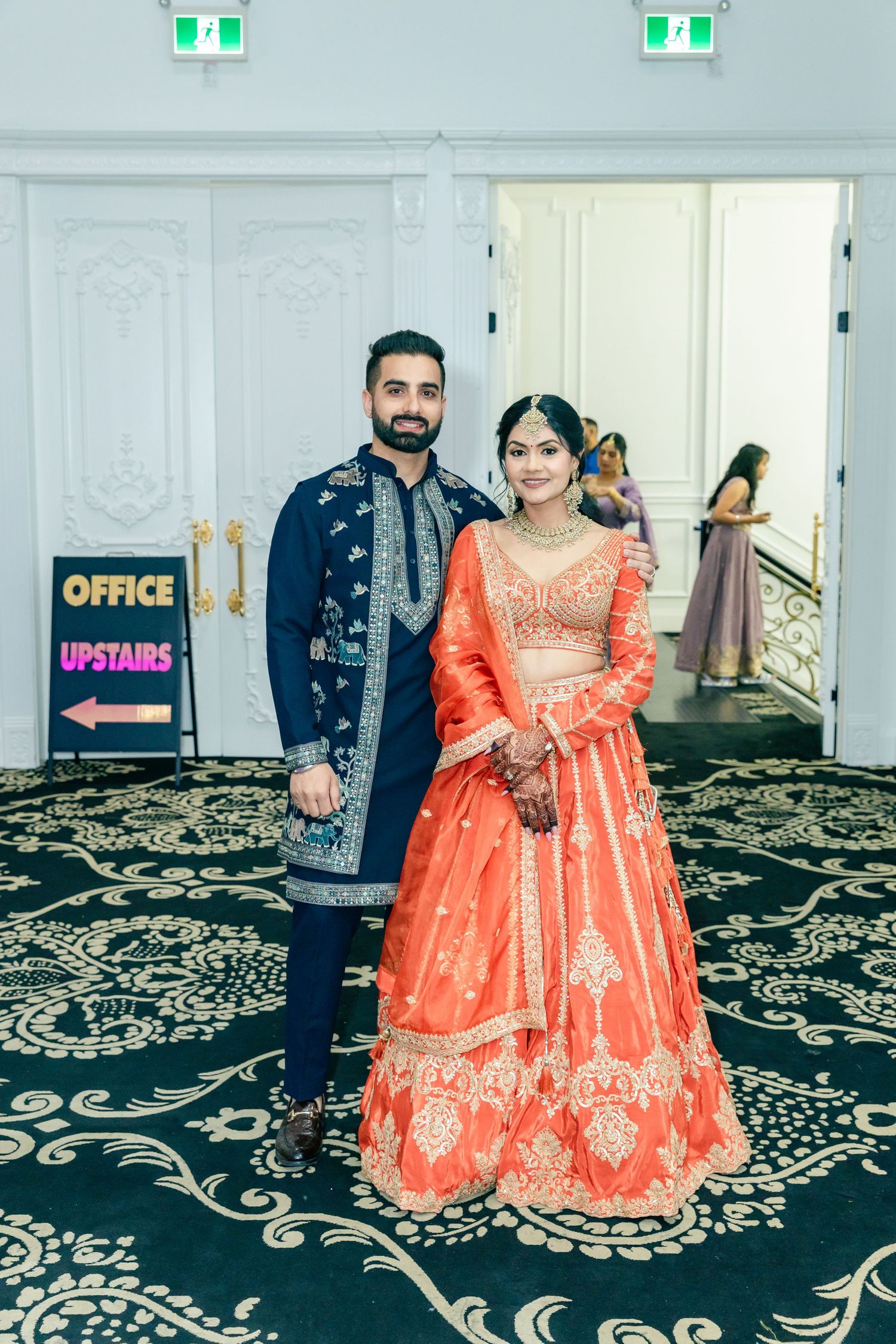 Couple in elegant Indian wedding attire posing on patterned carpet near office sign.