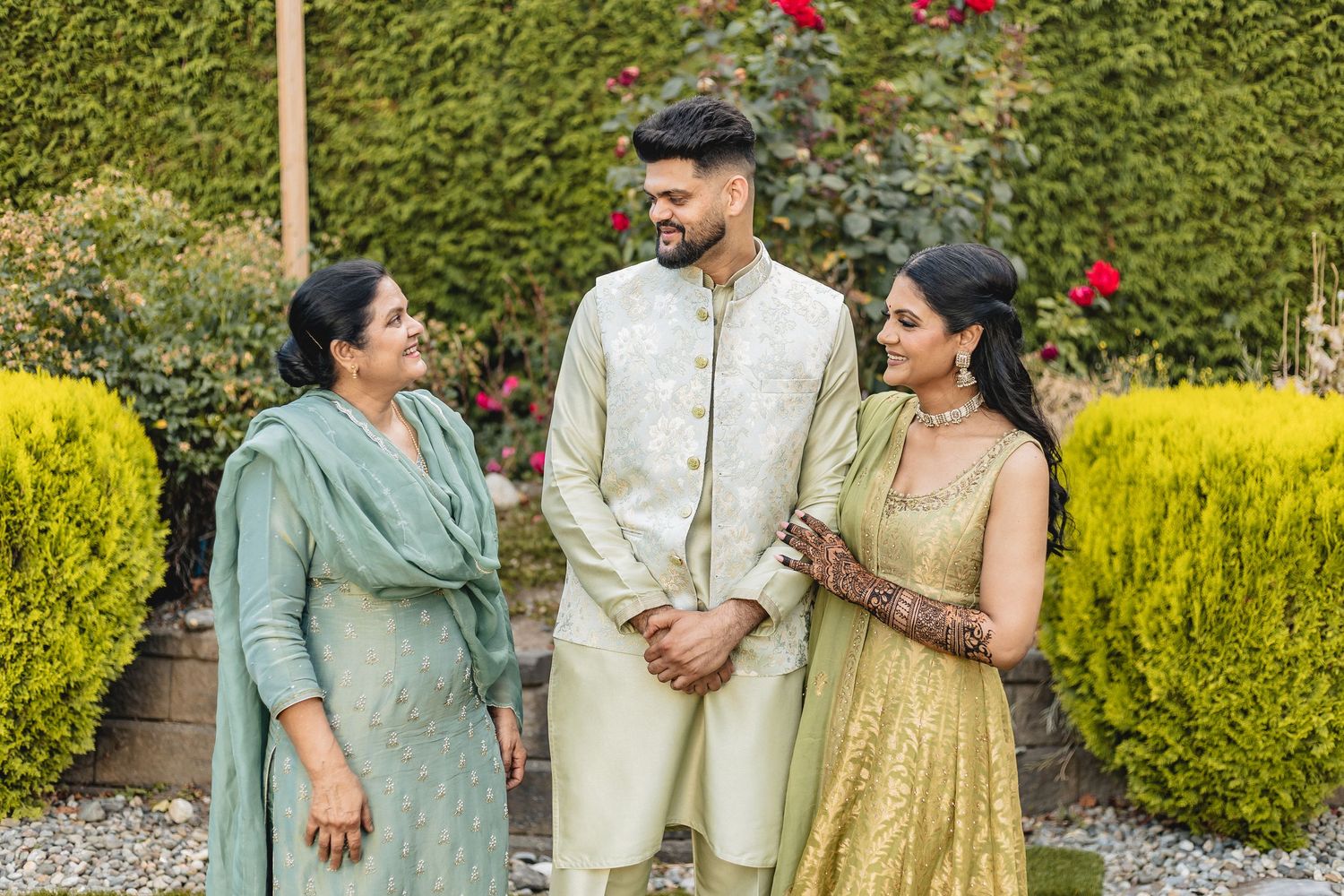 Couple poses together in traditional Indian attire against a lush garden backdrop with yellow and green foliage.