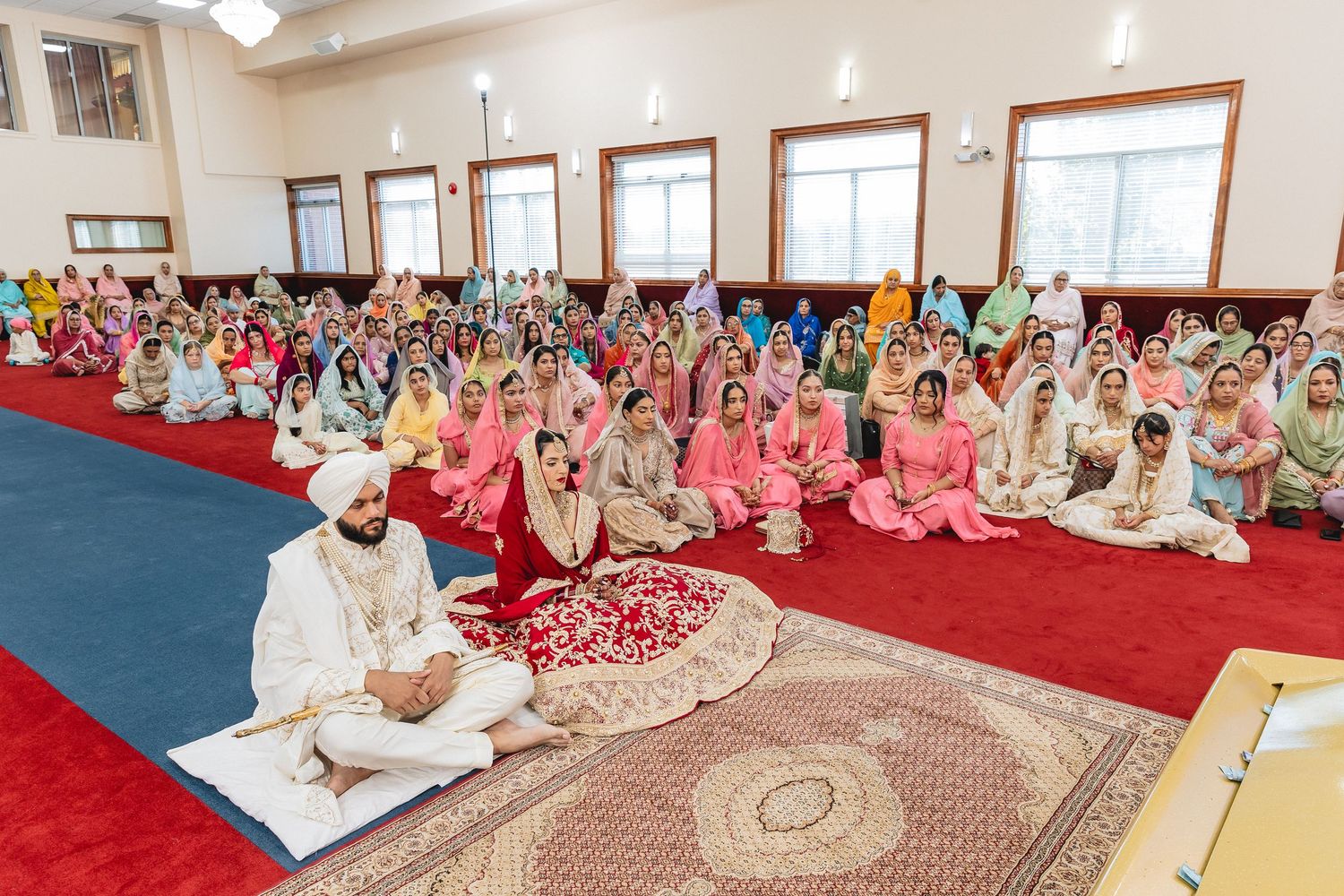 Large gathering of guests seated during Sikh wedding ceremony in decorated temple hall.