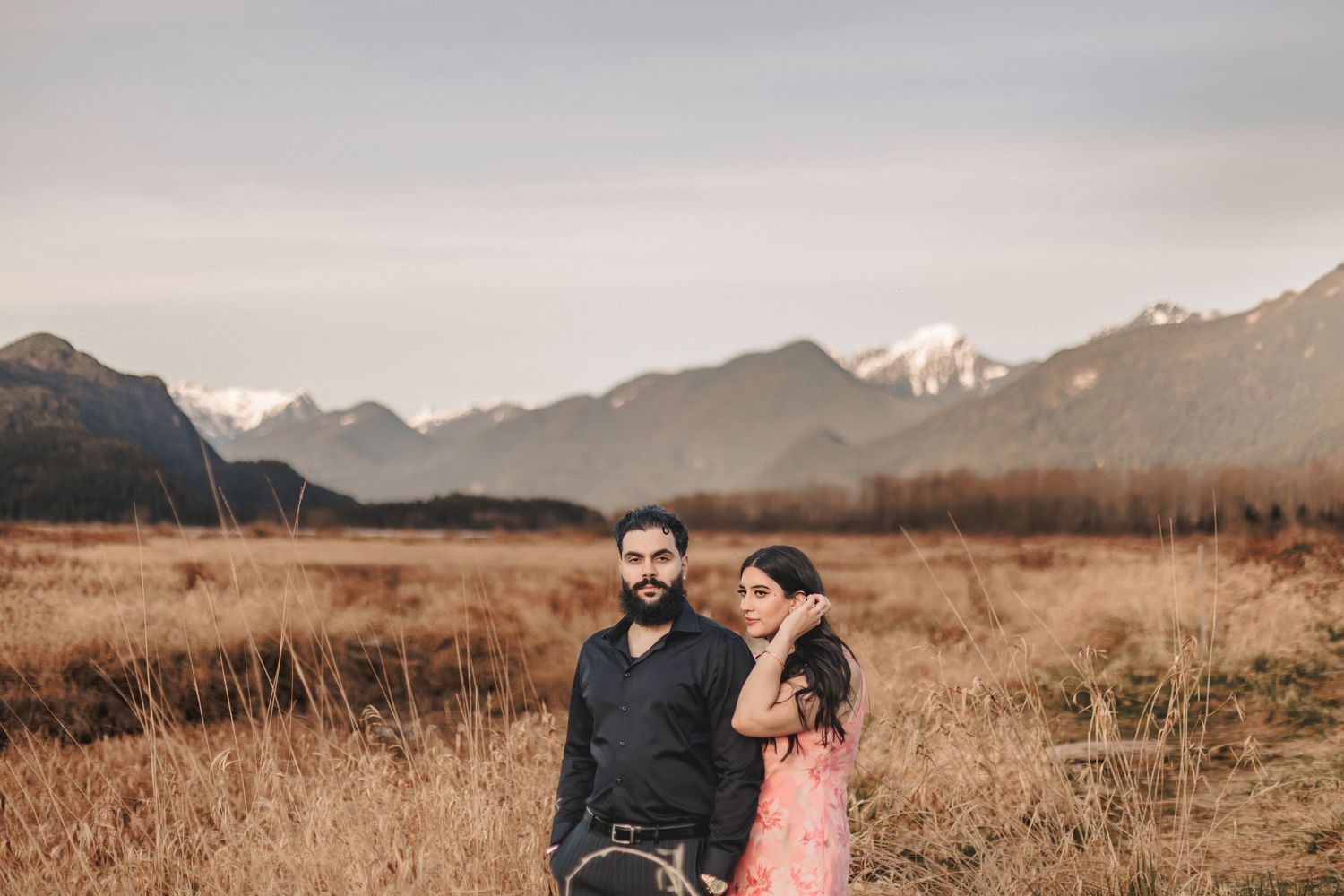 Couple poses together in a field with dramatic mountain peaks in the background during sunset.