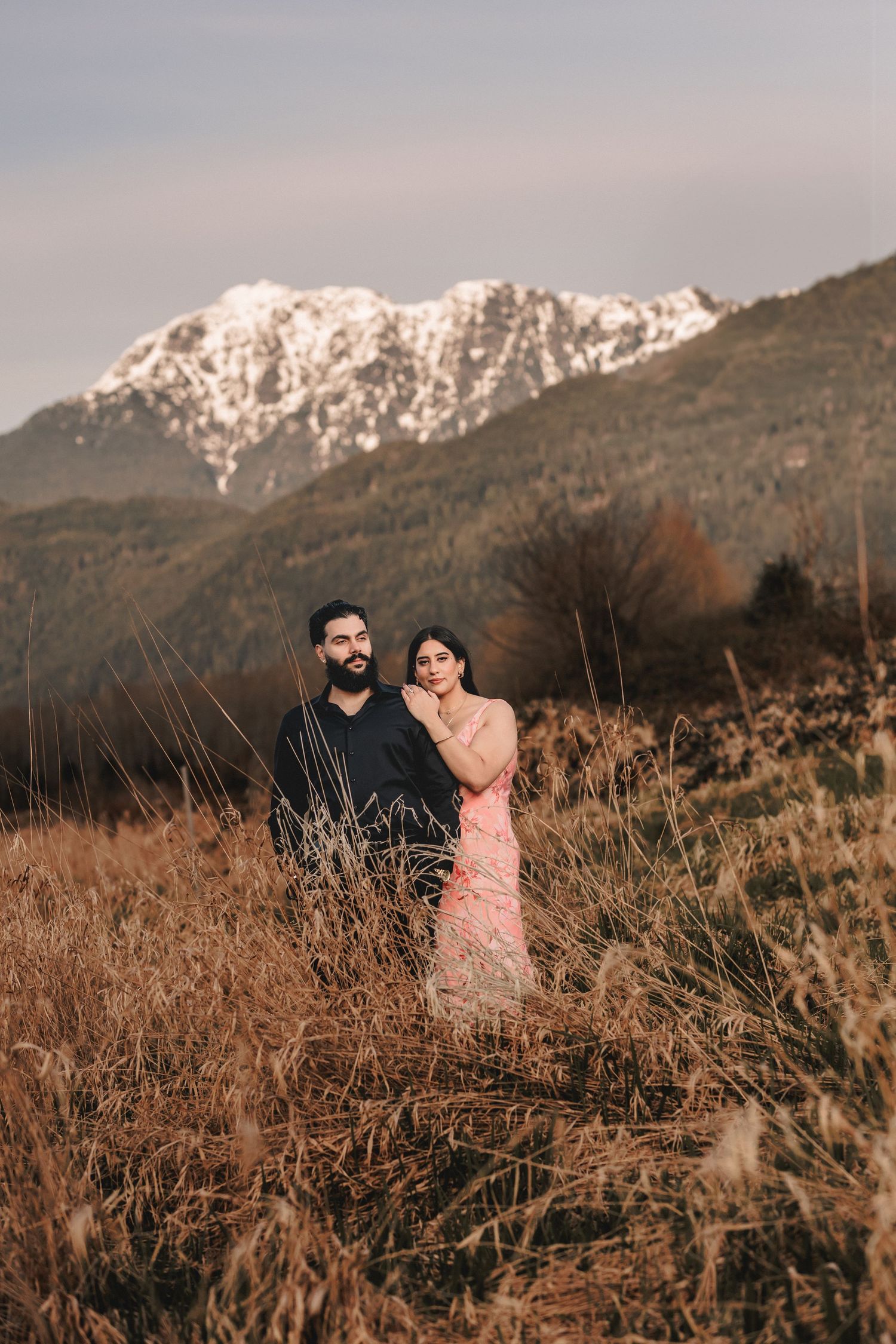 Couple embraces in a rustic field with majestic snow-covered mountains creating a dramatic backdrop. Pre Wedding Photoshoot in Vancouver