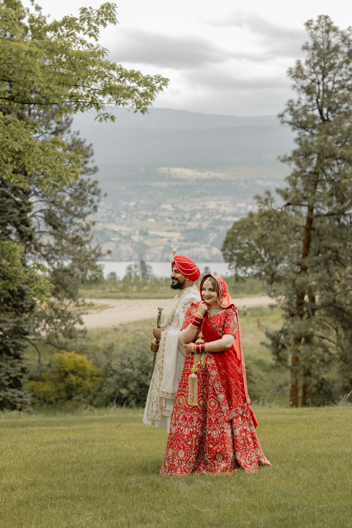 A couple in traditional Indian wedding attire embraces on a grassy hillside with mountains in the background.