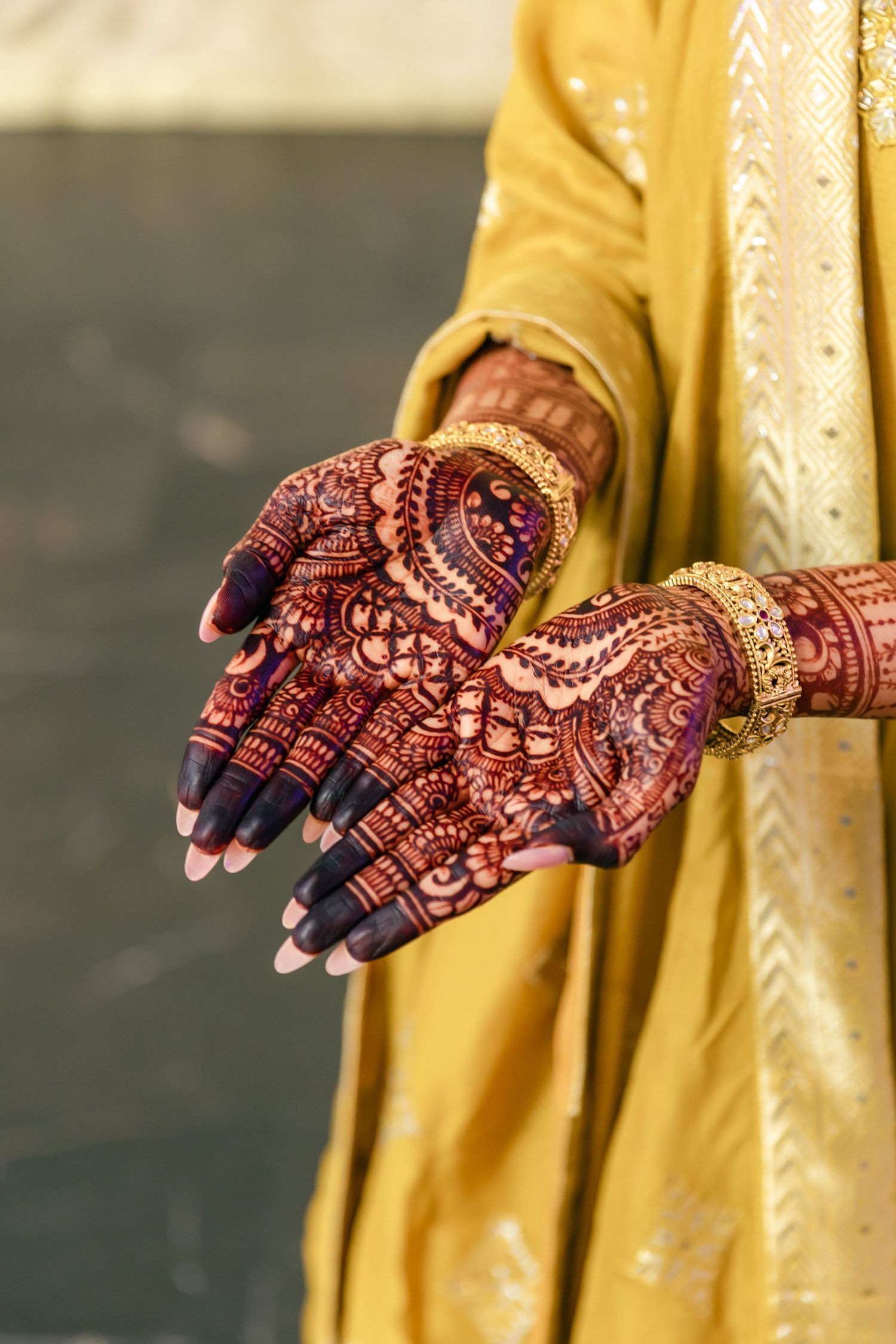 Detailed mehndi henna art patterns shown on hands wearing yellow traditional Indian attire.