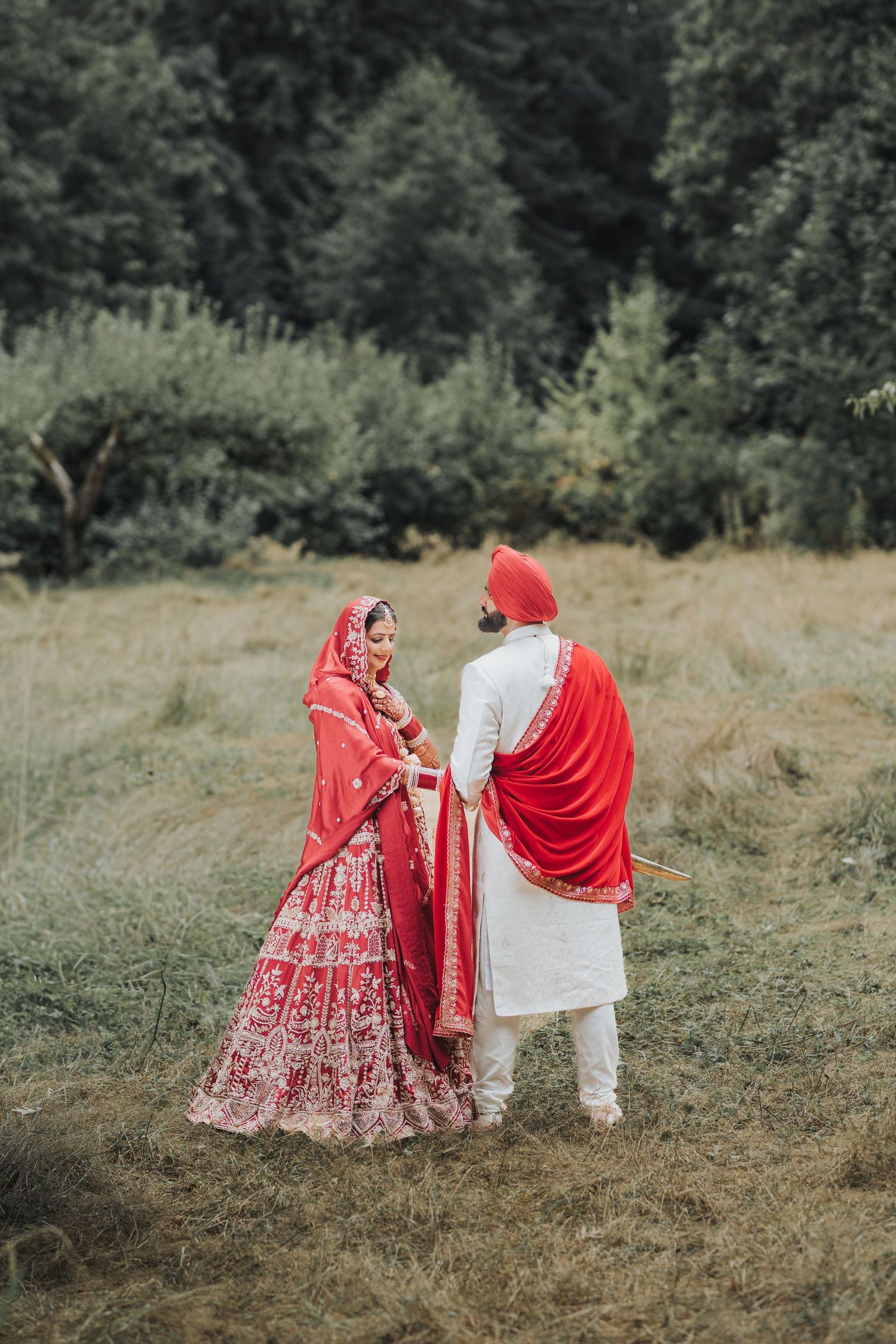 Wedding couple in red and white traditional Indian outfits posing in a natural outdoor setting.
