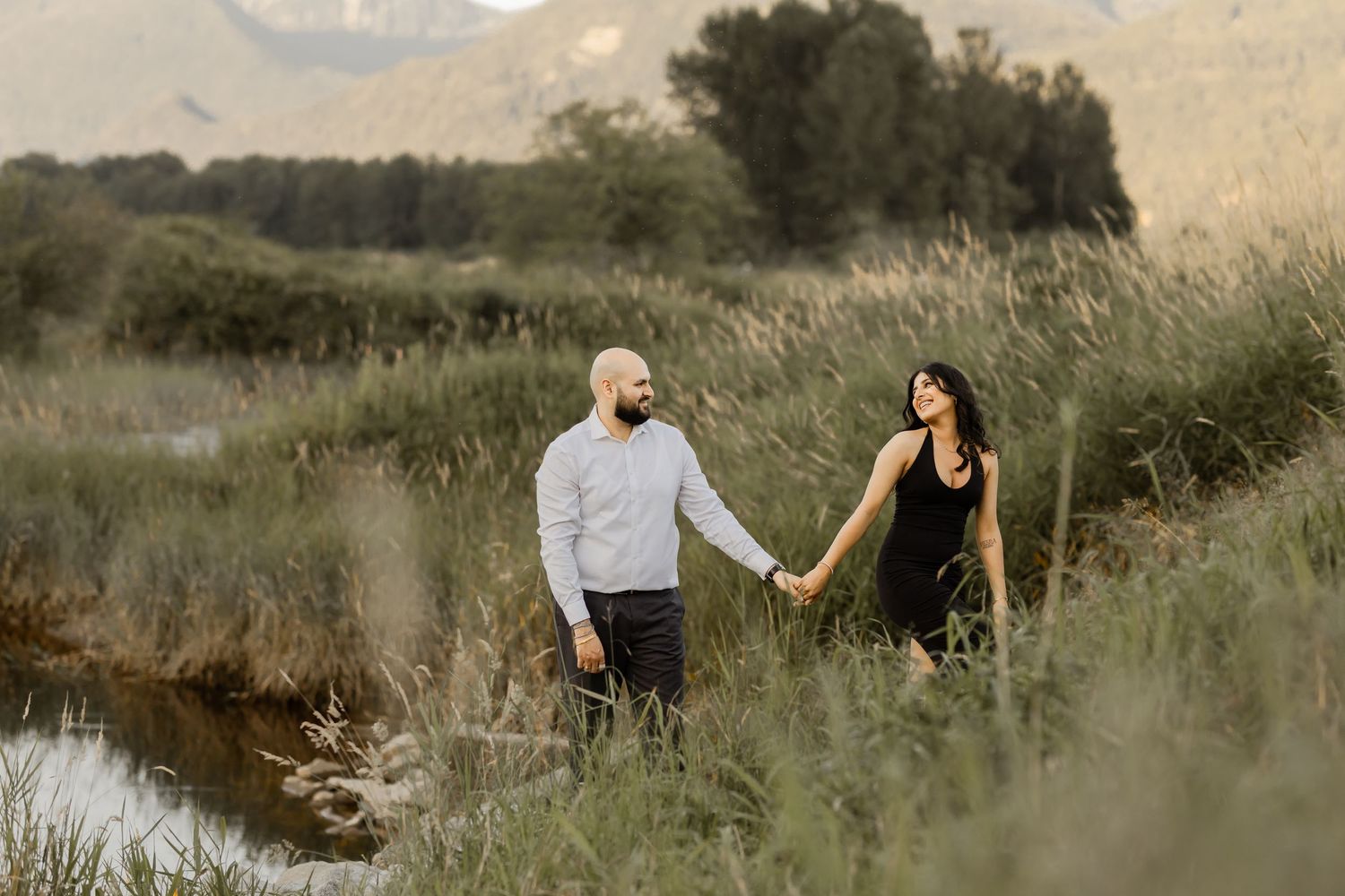 A couple walks hand in hand through tall grass at sunset.