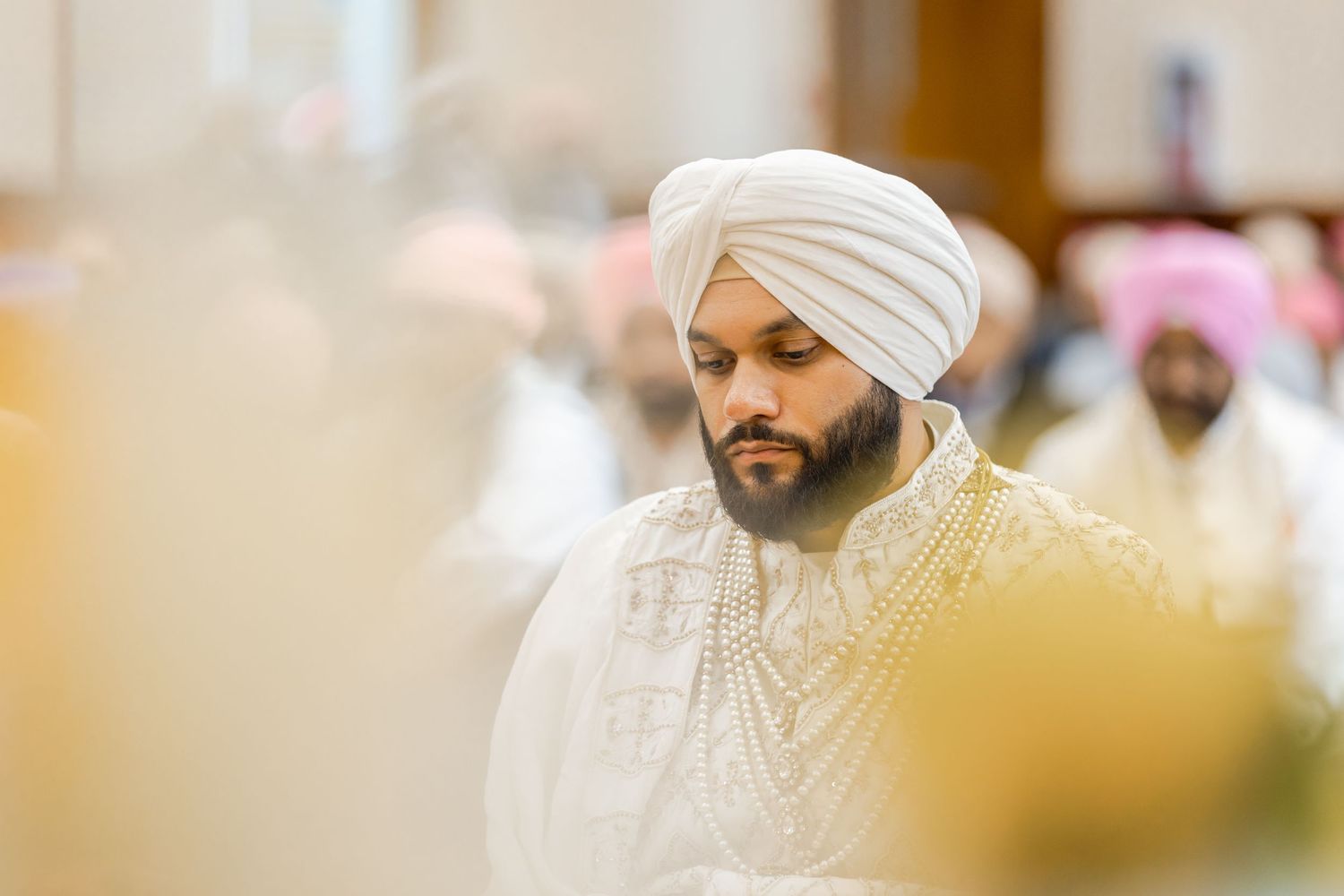 A person in traditional Sikh attire with white turban and embroidered sherwani sits during a ceremony.