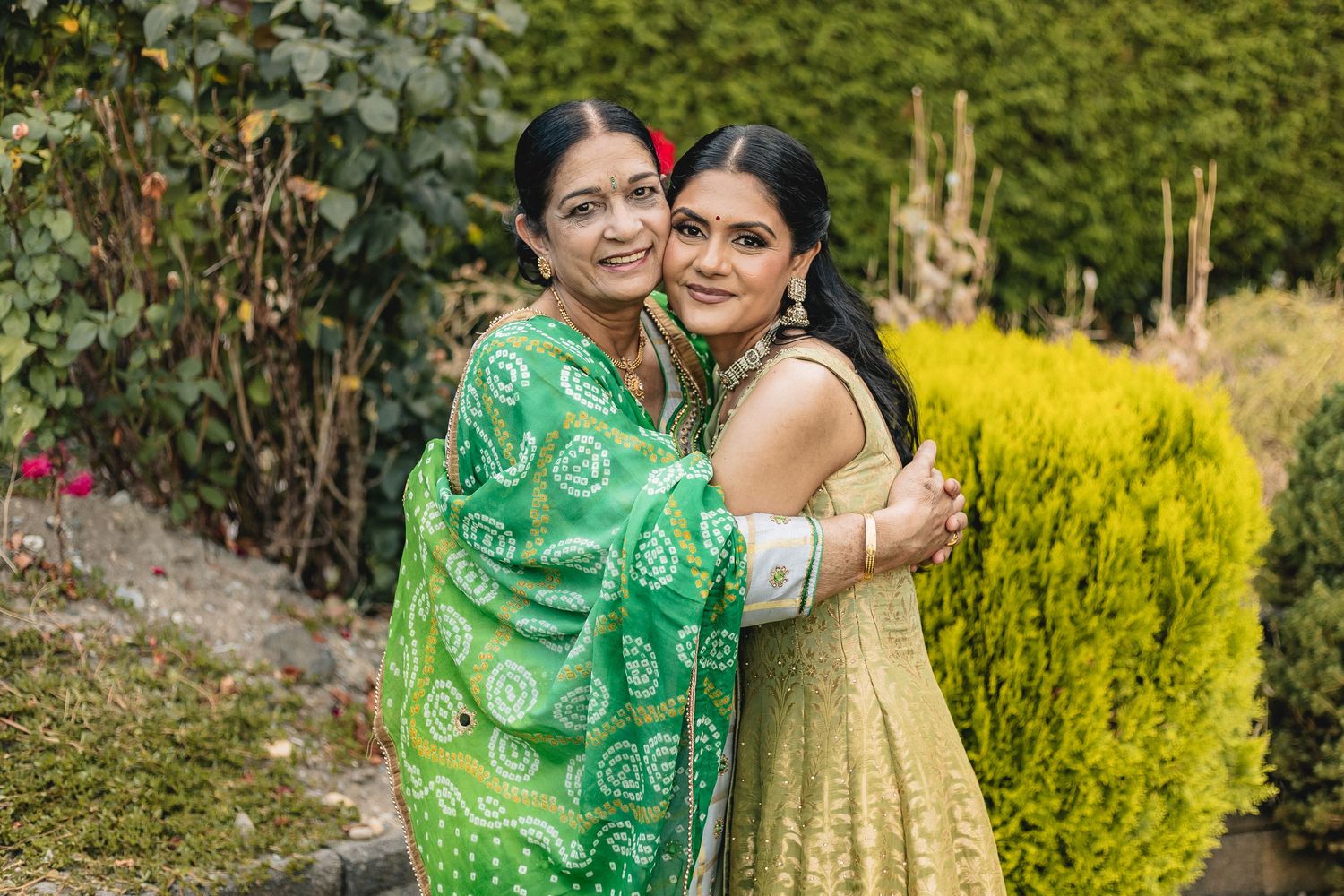 Couple in traditional Indian attire embracing outdoors with green foliage and yellow flowers in background.