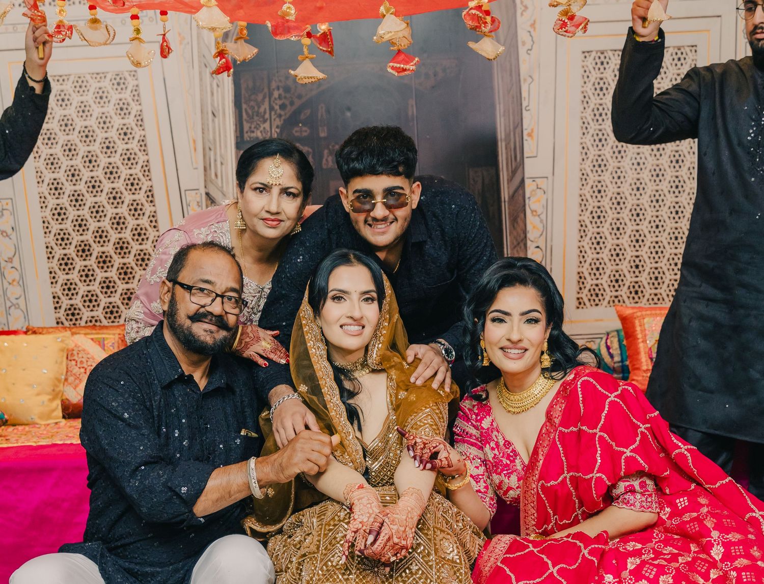 A group celebrates at a festive Indian wedding reception with bright red and gold decorations in the background.