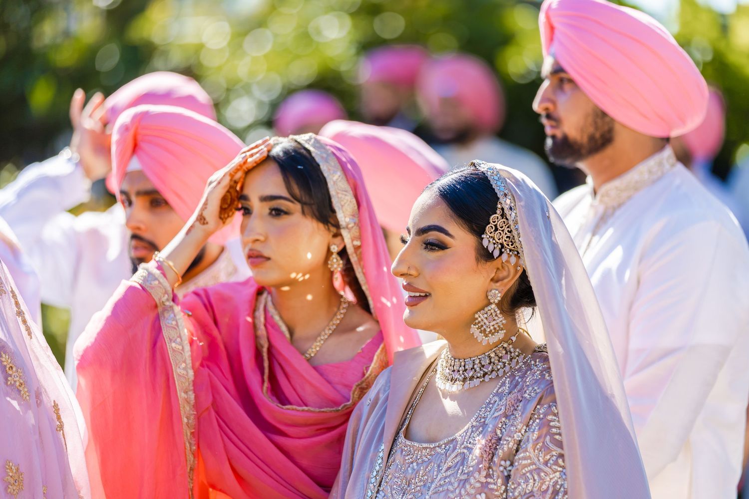 Wedding guests in traditional Punjabi attire with pink turbans and dupattas gather outdoors in sunlight.