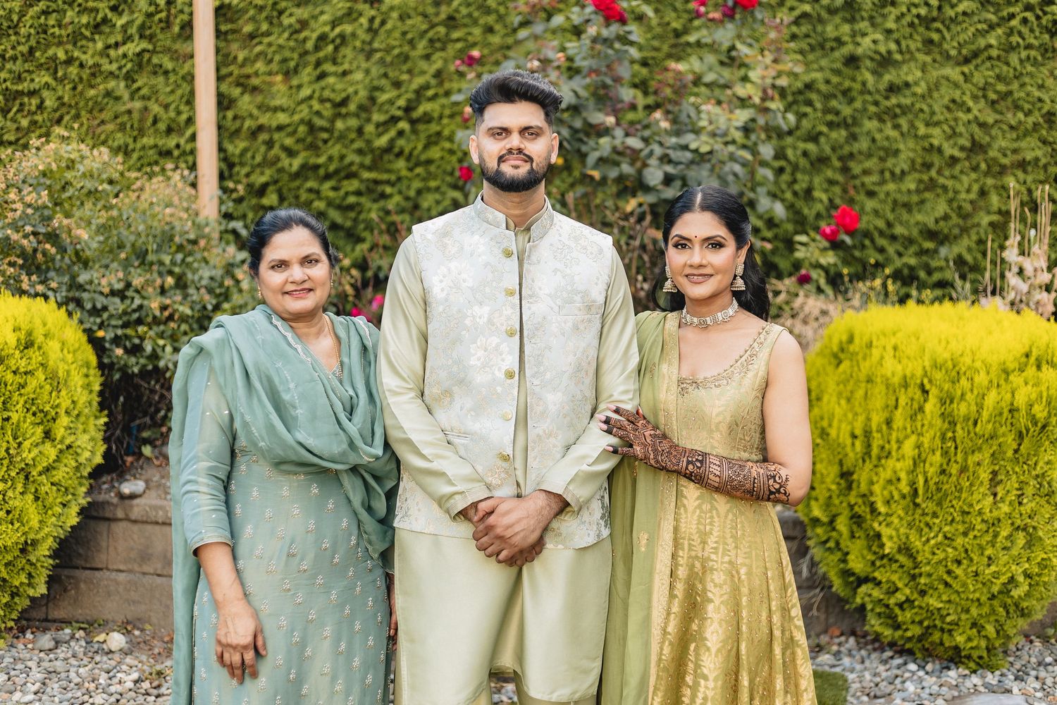 A group in traditional Indian attire poses together in a garden setting with red flowers and green bushes in the background.