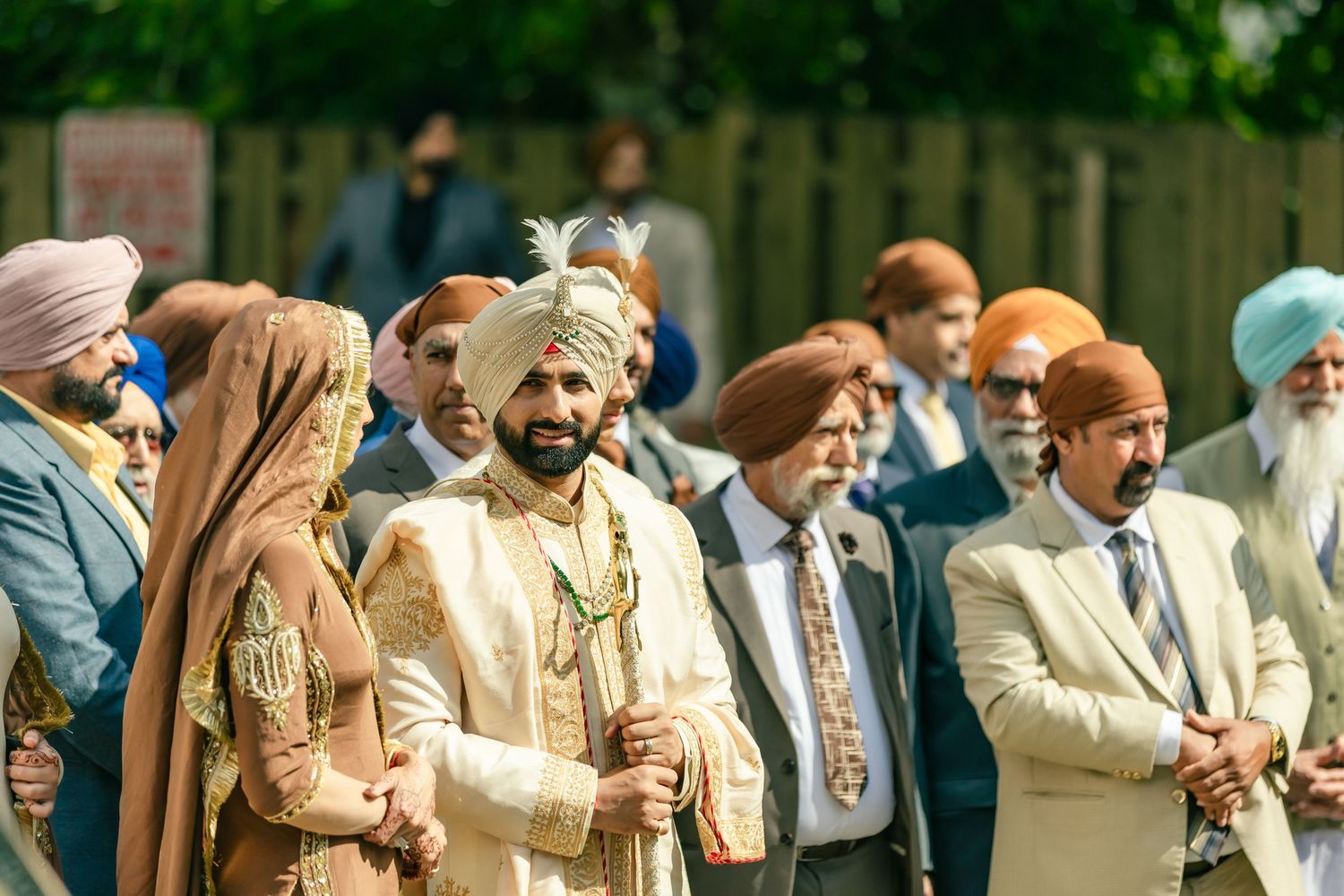 A line of guests in traditional formal wear stand together at an outdoor event.
