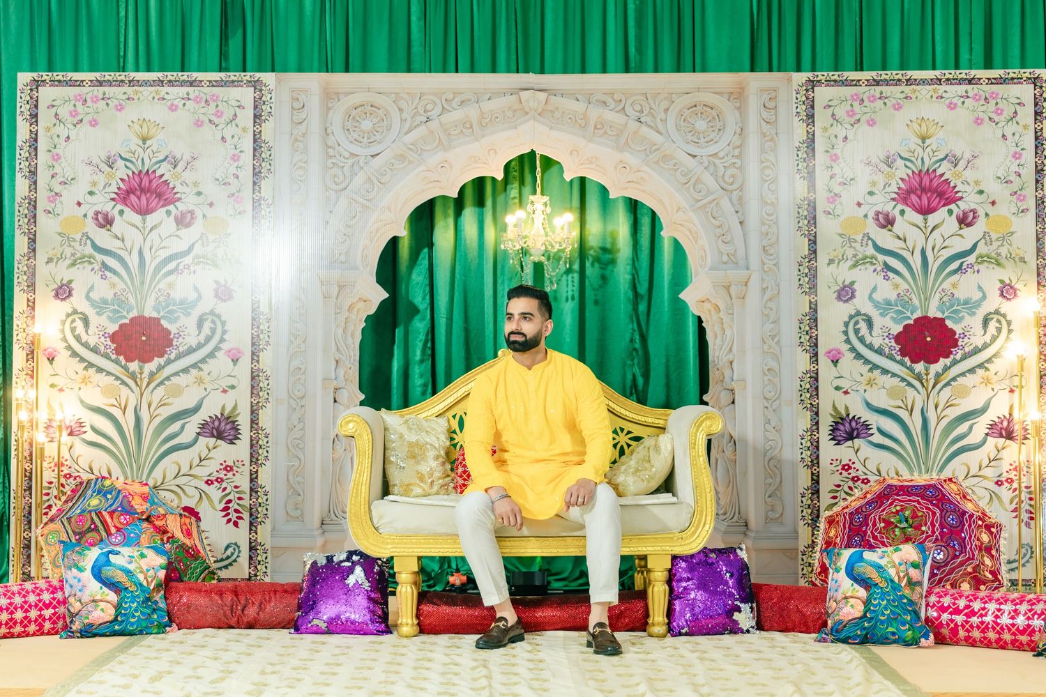 Person in yellow attire seated on decorative throne with green and floral backdrop.