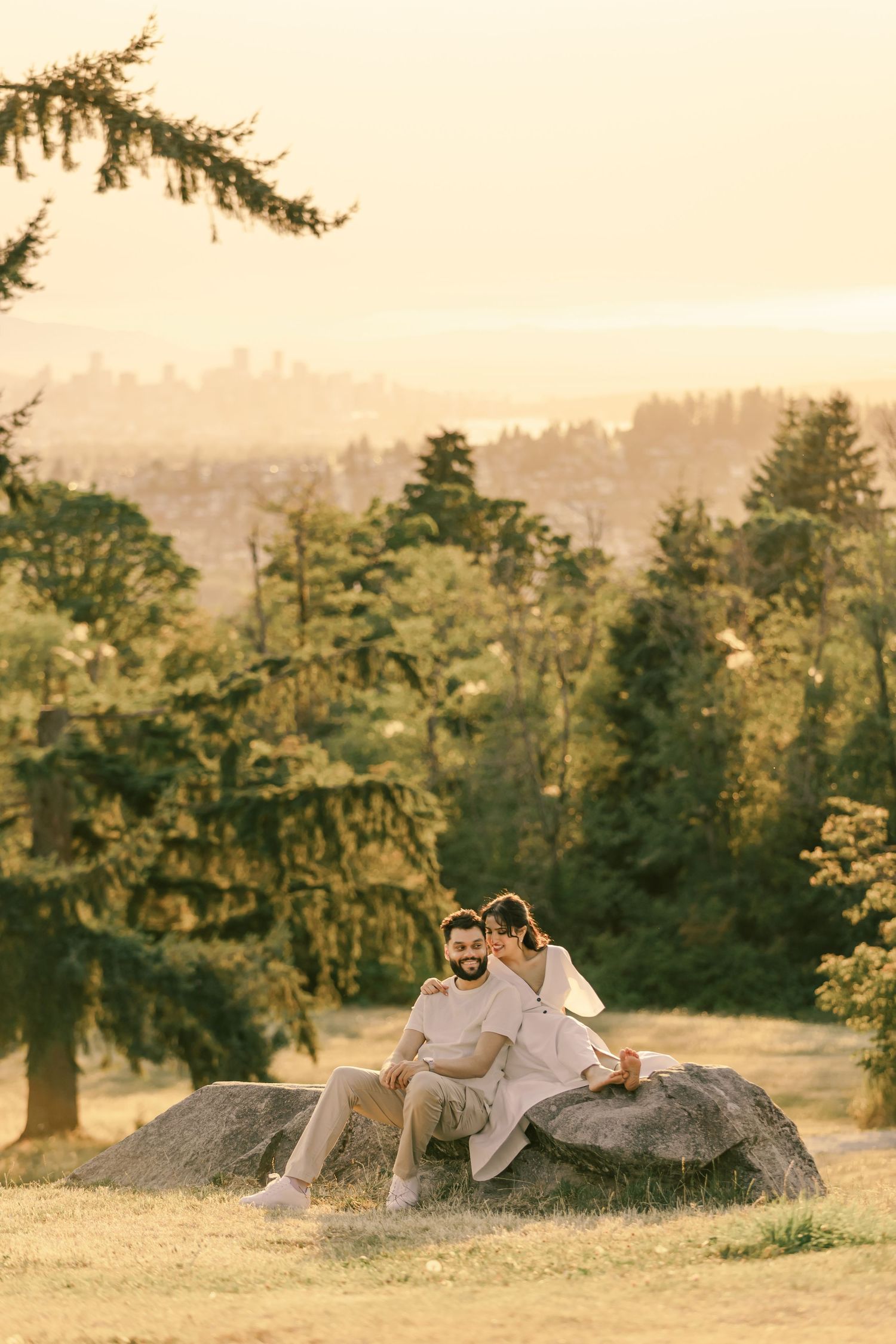 Two people in light-colored outfits relax on a large rock overlooking a scenic mountain landscape.
