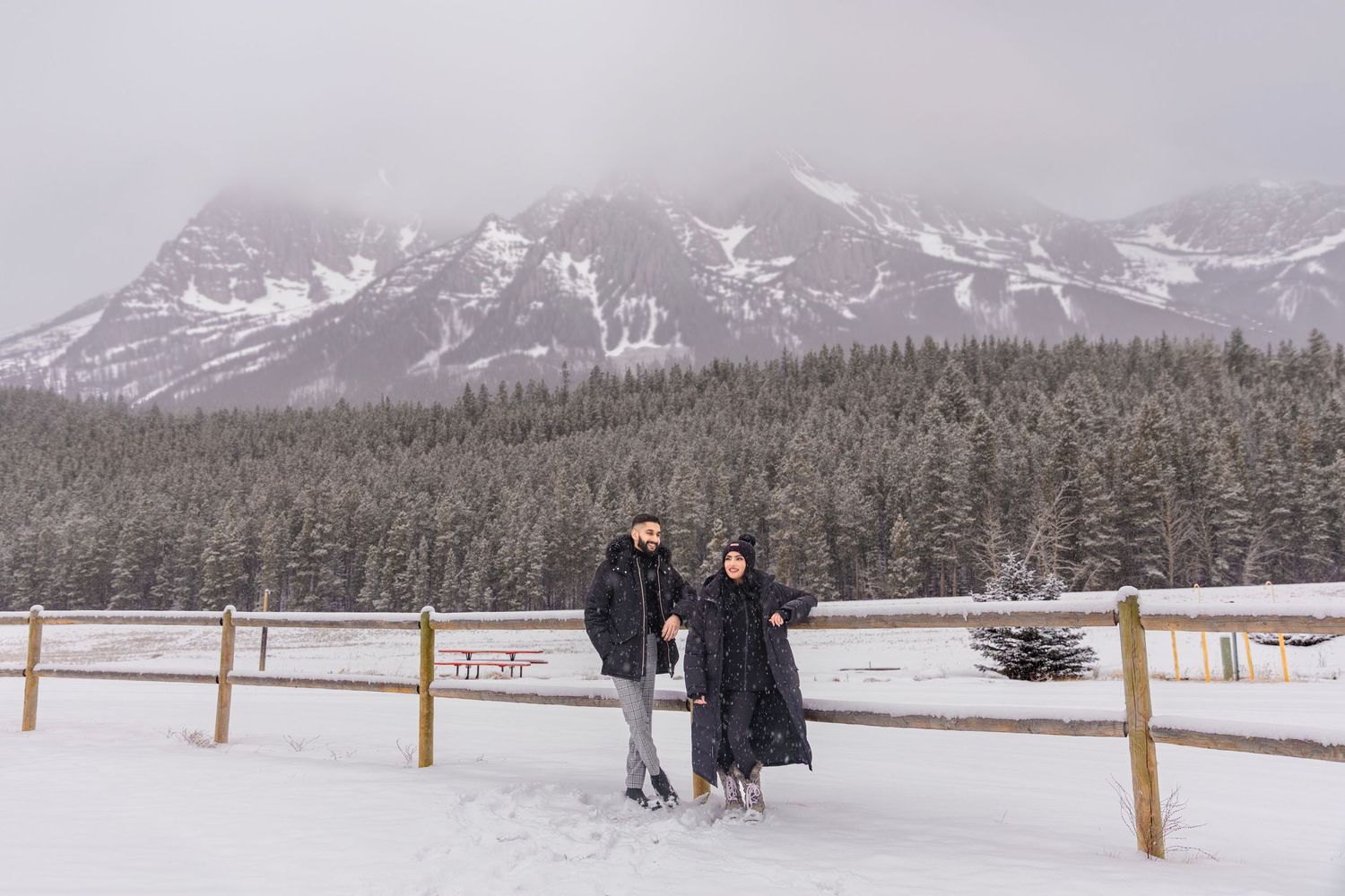 Couple standing by wooden fence in snowy mountain landscape with evergreen forest and misty peaks in winter.