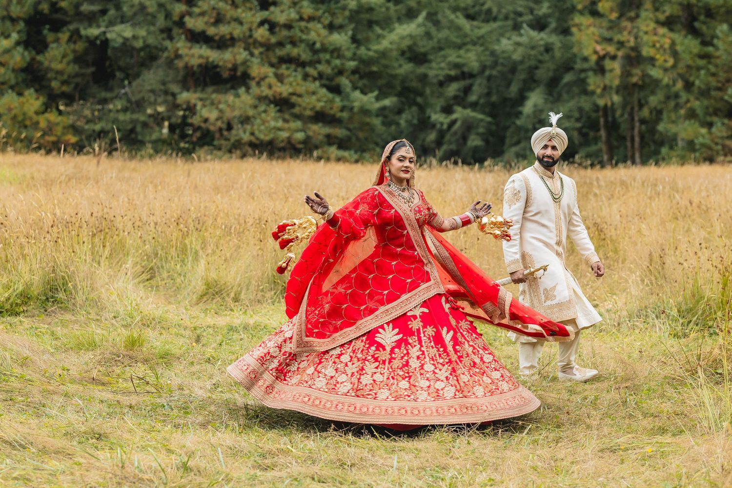 A couple in traditional Indian wedding attire posing in a sunlit meadow with tall grass.