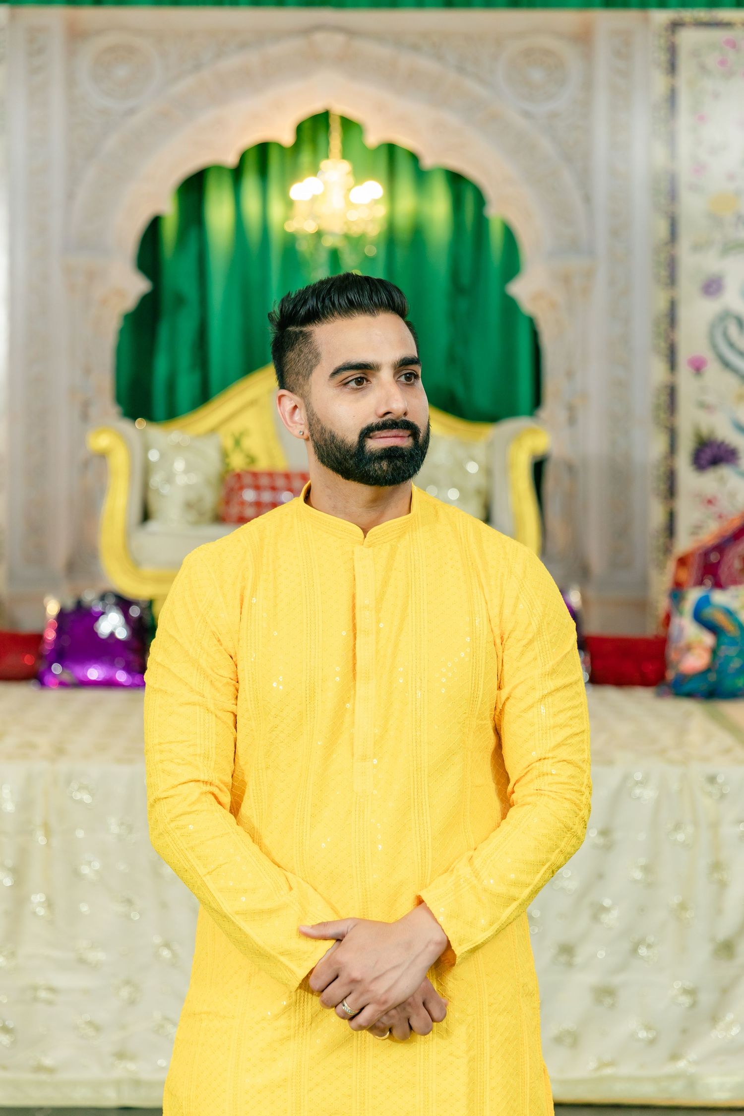 A person wearing bright yellow traditional attire poses in front of an ornate green and white backdrop.
