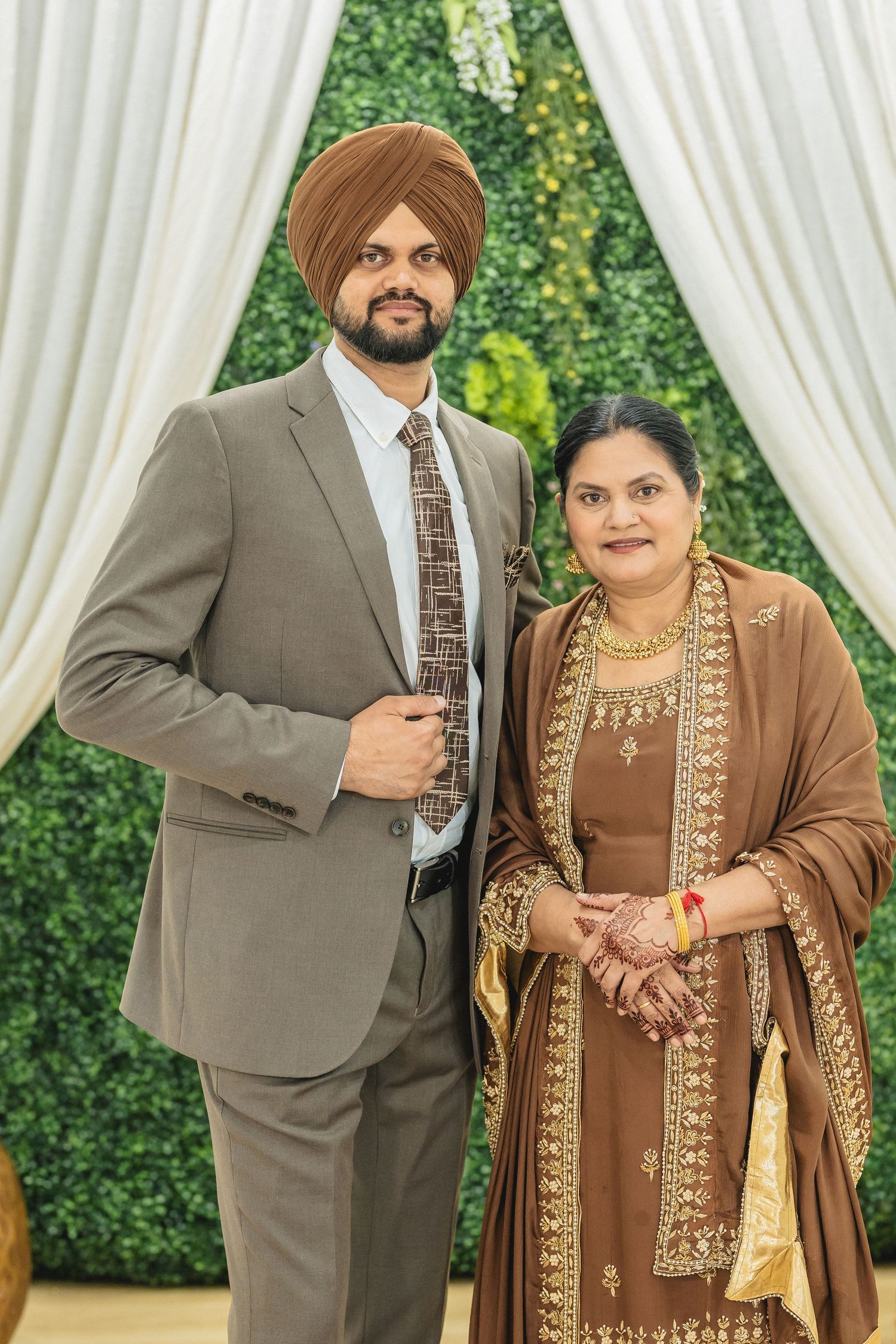 Traditional Indian wedding guests in formal attire stand together against a green hedge and white curtain backdrop.