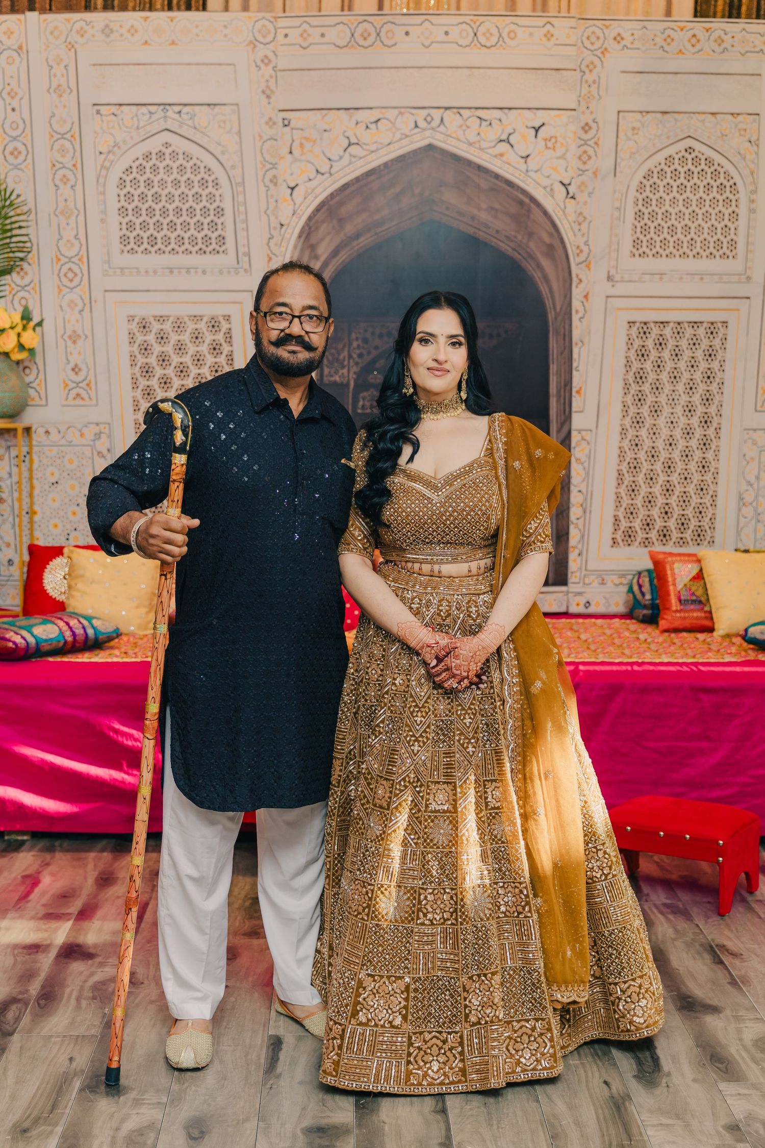 Couple wearing traditional South Asian wedding attire standing against an intricately decorated backdrop.