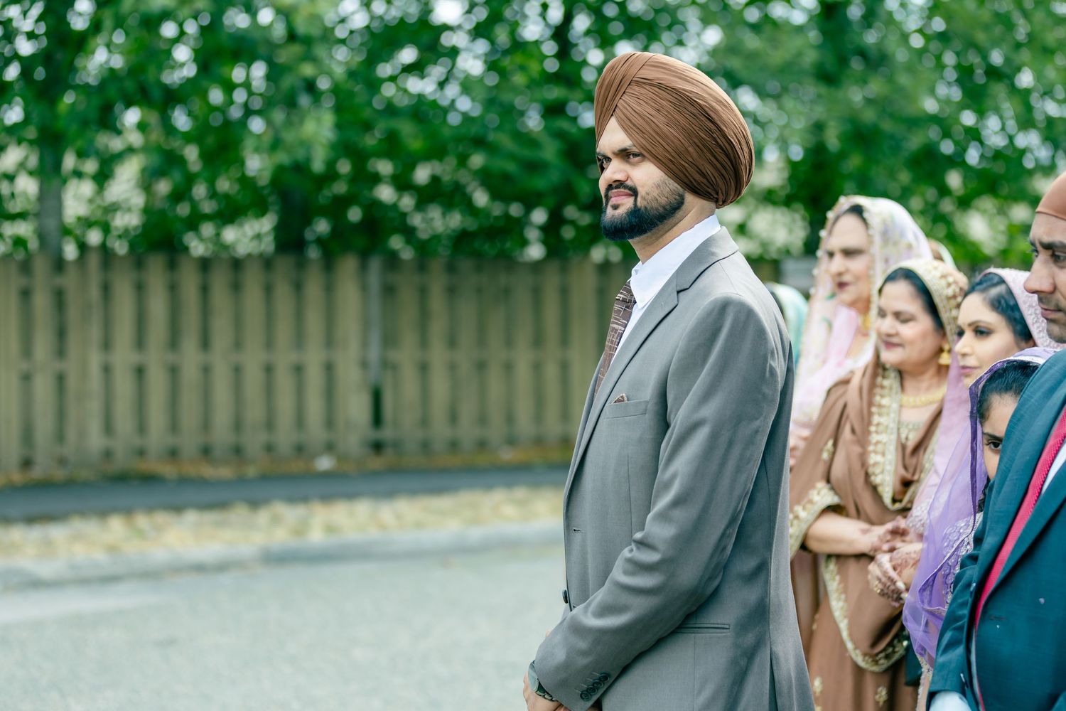 Wedding guests in formal attire gather outdoors on a sunny day surrounded by green foliage.