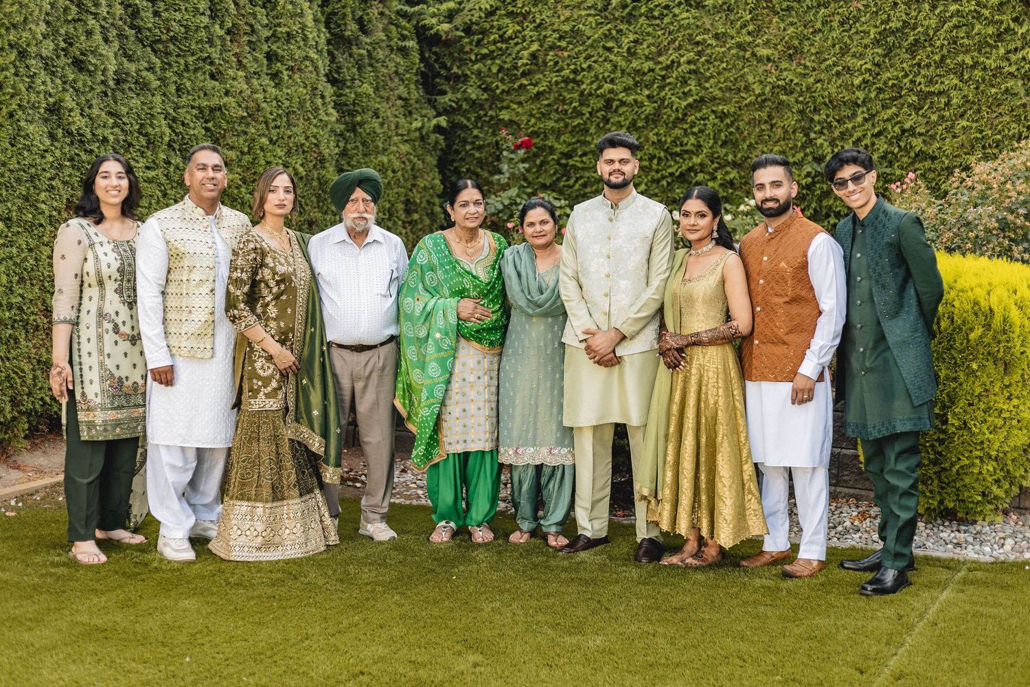 A group in traditional South Asian attire poses together in a garden with green hedges and yellow flowers.