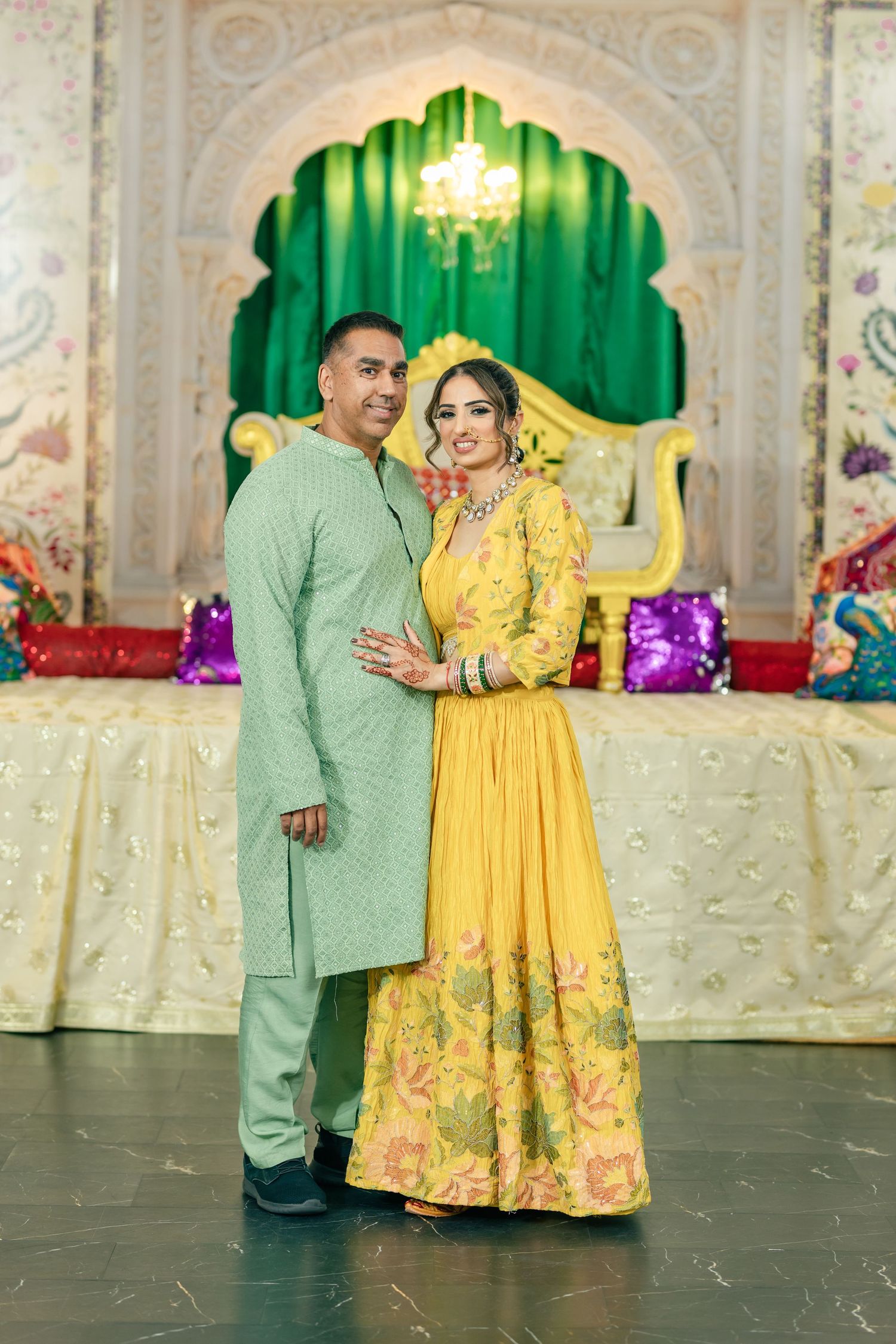 A couple in traditional attire poses together in a decorated venue with green and yellow decor elements.