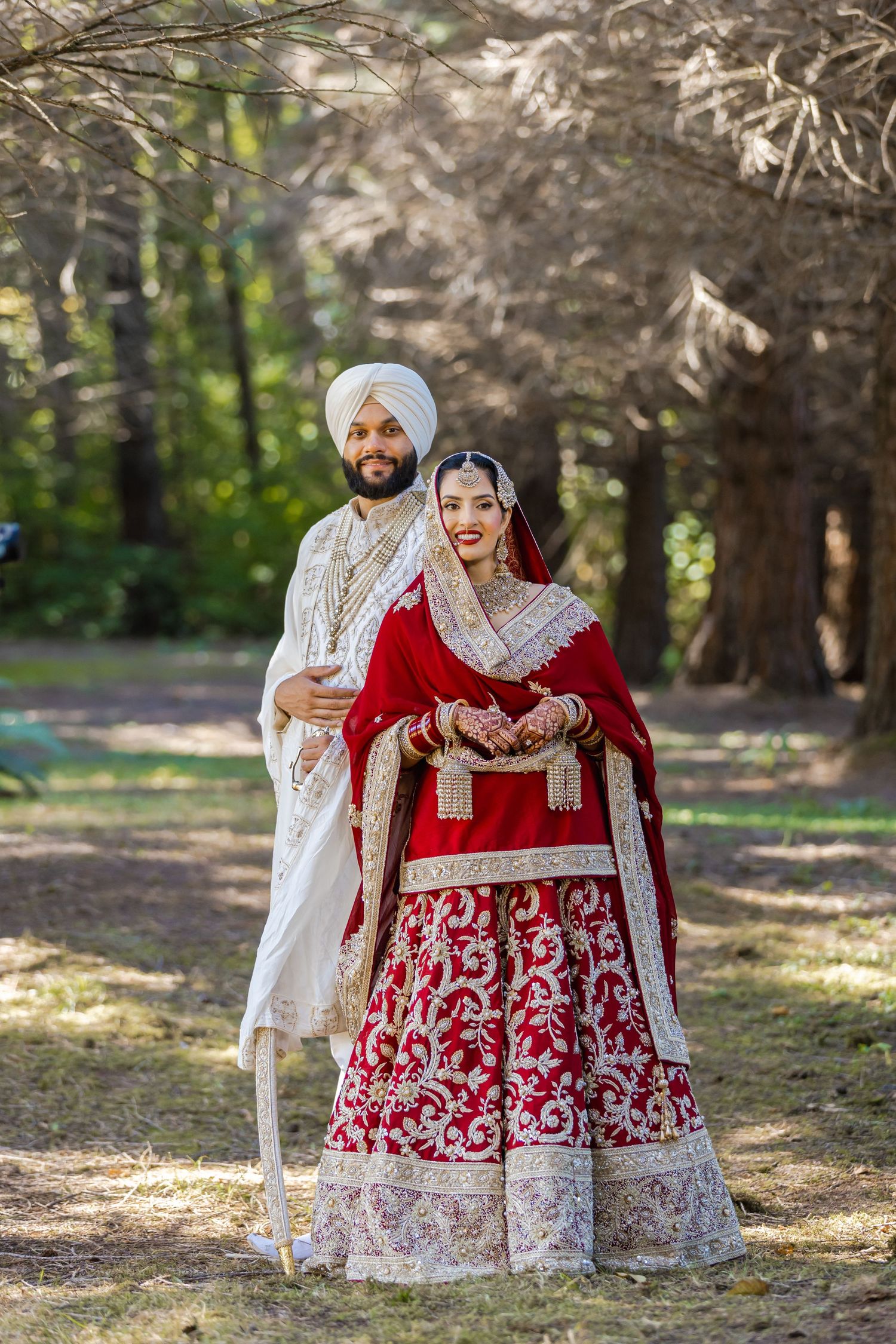 A couple in traditional Sikh wedding attire poses under cherry blossom trees in a park setting.