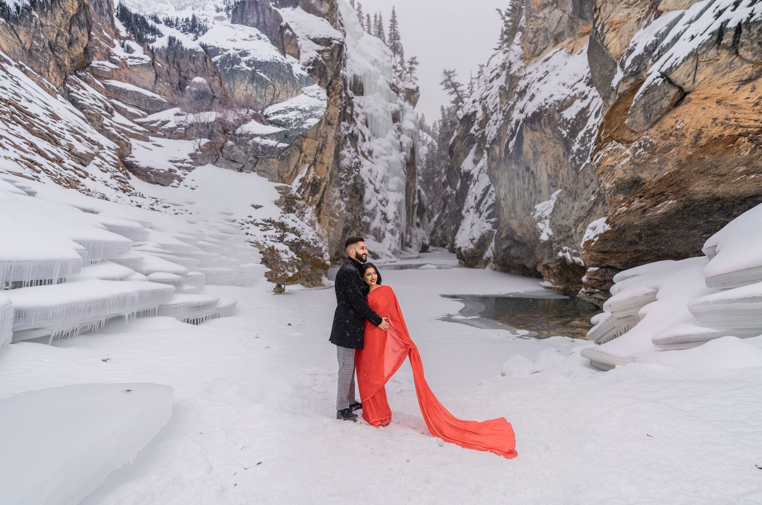 Couple embracing in snowy canyon landscape, woman in flowing red dress, romantic winter engagement photo session.