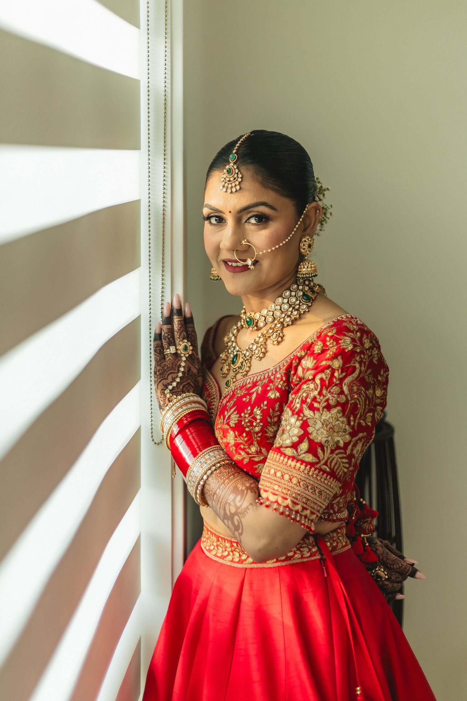Traditional Indian bride in red lehenga with gold jewelry poses by window with henna decorated hands.
